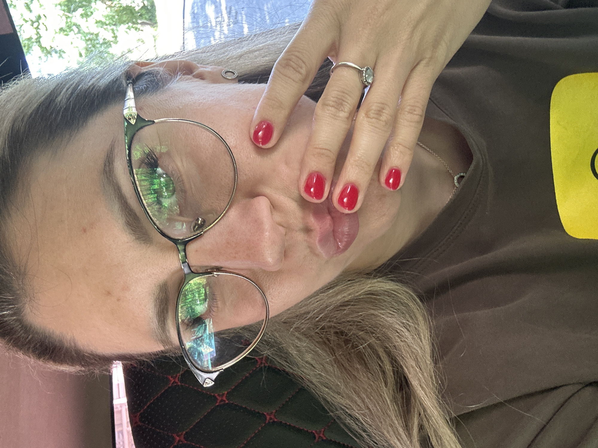 This image is a selfie taken by a woman in Novoaltaysk, Russia. She is wearing glasses and a brown t-shirt, and her fingernails are painted red. Her hand is raised to her face, and she appears to be making a kissing gesture. The background is blurred but appears to be outdoors, with trees and a vehicle visible.