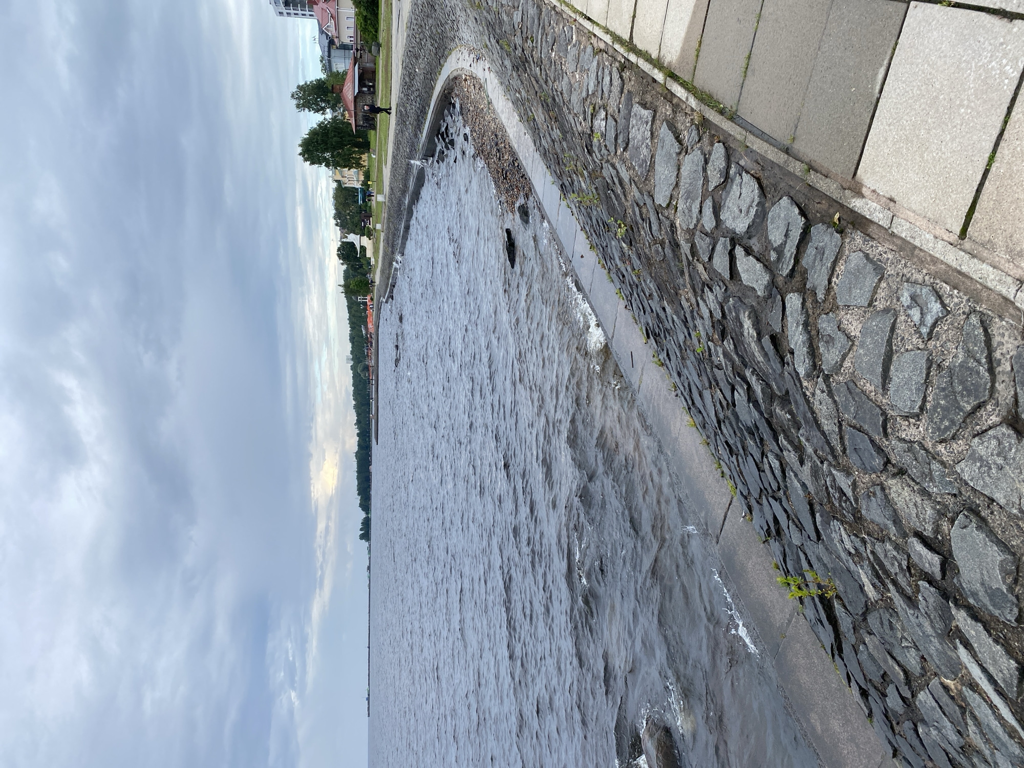A paved embankment lines the shore of a large body of water under a cloudy sky. The embankment is constructed of stone slabs at the water's edge and transitions to a lighter-colored paved walkway further inland. A single person walks along the paved walkway. In the background, buildings and trees are visible along the distant shoreline. This scene is located in Petrozavodsk, Russia, on the shore of Lake Onega.