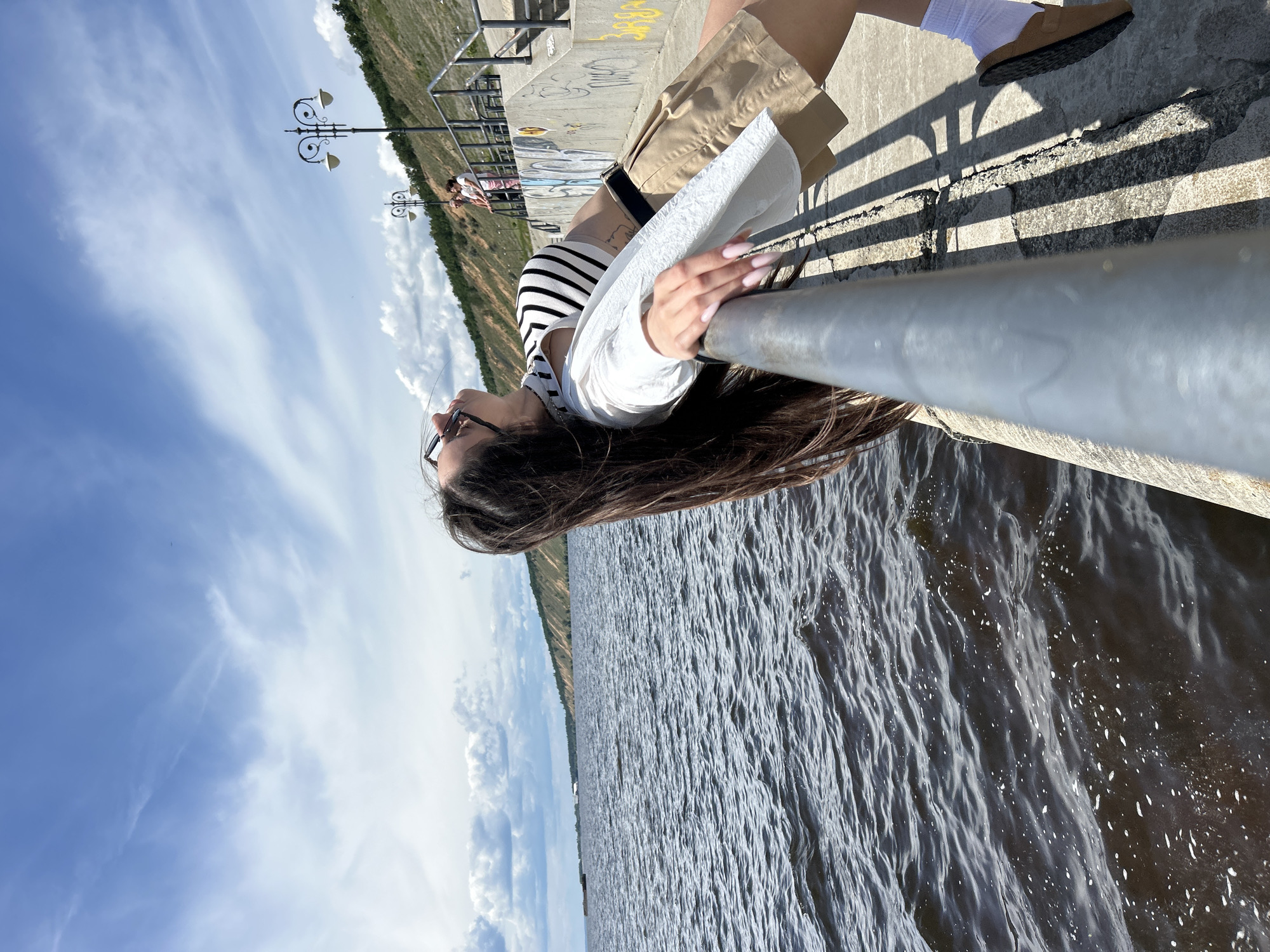 A young woman with long, dark hair is captured in a dynamic pose, leaning back against a metal railing with her head tilted upwards towards a bright blue sky filled with scattered clouds. She is wearing a black and white striped crop top, a white open shirt, beige shorts, white socks, and brown sandals. Her gaze is directed away from the camera, suggesting a moment of relaxation or contemplation.
The scene appears to be a waterfront promenade, likely in the city of Tolyatti, Russia. The railing she is leaning on is positioned at the edge of a concrete embankment. Below the embankment, dark, choppy water stretches out, reflecting the sky. In the background, a steep, vegetated hillside rises, dotted with trees and featuring a prominent sandy or rocky cliff face. A pathway with a railing and lampposts runs along the top of the embankment, and there are hints of graffiti on the concrete structures. Two other individuals are visible in the distance on the pathway, adding a sense of a public space. The overall impression is one of a sunny, breezy day by a body of water.