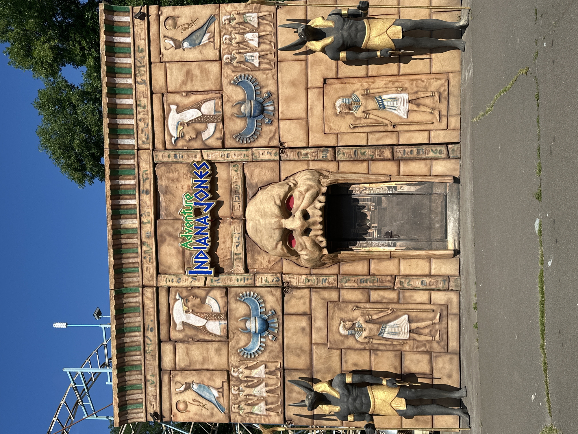 The image shows the entrance to an attraction called "Adventure Indiana Jones," designed to resemble an Egyptian temple. The facade is made of tan-colored blocks, intricately carved with hieroglyphics and figures of pharaohs and birds. Two large statues of Anubis, the jackal-headed god, stand on either side of the entrance. The entrance itself is a large, menacing, carved face with glowing red eyes and sharp teeth, serving as a doorway. The sky is clear and blue, and green trees are visible behind the structure. A roller coaster track is partially visible to the left of the facade. The ground in front of the attraction is paved with asphalt and has patches of grass growing through cracks.