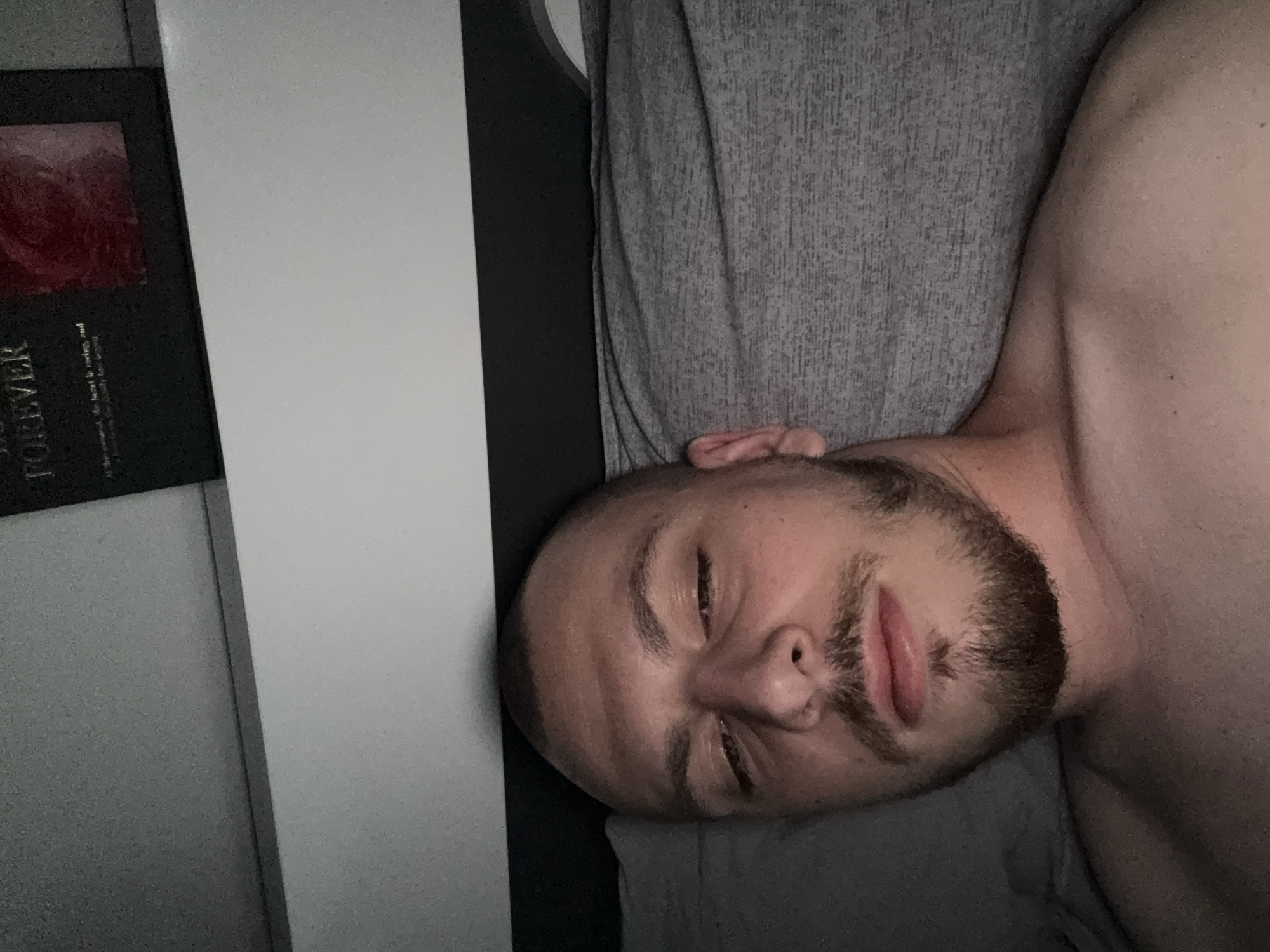 The image is a close-up, overhead shot of a man lying on his back in bed. He has a shaved head, light skin, and a beard and mustache. His eyes are partially closed, and he is looking upwards and slightly to his right. He is shirtless, wearing a gray t-shirt that is visible on his shoulders and chest. The bed appears to be made with a gray textured comforter or duvet. In the background, a black object with gold lettering that reads "FOREVER" is partially visible, along with a framed image of a red rose. The lighting suggests it is nighttime or the room is dimly lit. The scene takes place indoors, likely in a bedroom. There is no indication of the specific location in Stara Zagora, Bulgaria, within the image itself.