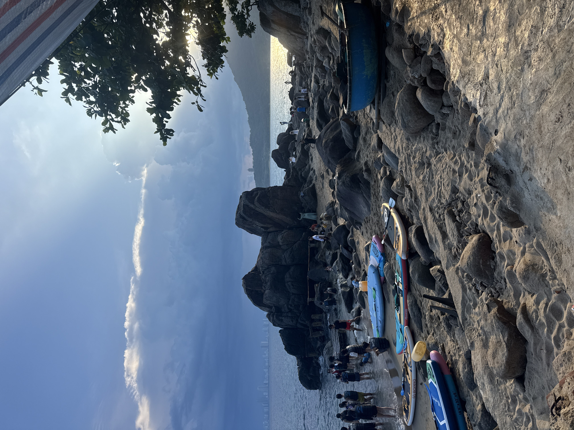 The image depicts a crowded beach scene in the Unknown city of Unknown. A group of people is gathered at the shoreline, near a rocky outcrop. Several paddleboards are lying on the rocky beach. A blue boat sits near the top of the rocky area. A tree hangs over the scene from the top left. In the background, the sea meets the horizon under a cloudy sky with the sun barely peaking through. Tall buildings appear faintly in the distance.