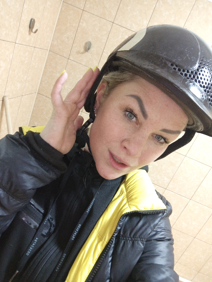 A female individual is depicted in a close-up selfie, captured at a high angle in an indoor setting. She wears a dark helmet with a white stripe and perforated ventilation. Her right hand, featuring yellow-painted fingernails, is positioned at the helmet's side, appearing to adjust it. Her face shows blue eyes, long eyelashes, defined eyebrows, and freckles across fair skin, with her mouth slightly parted. She is dressed in a black and yellow puffer jacket, zipped, over a dark garment with a visible black strap bearing the text "002829100" and "ABTACADO". The background reveals a light beige tiled wall with two metallic wall hooks and a light-colored wooden dowel visible vertically in the lower-left corner. This scene is located in the city of Gorka, Russia.