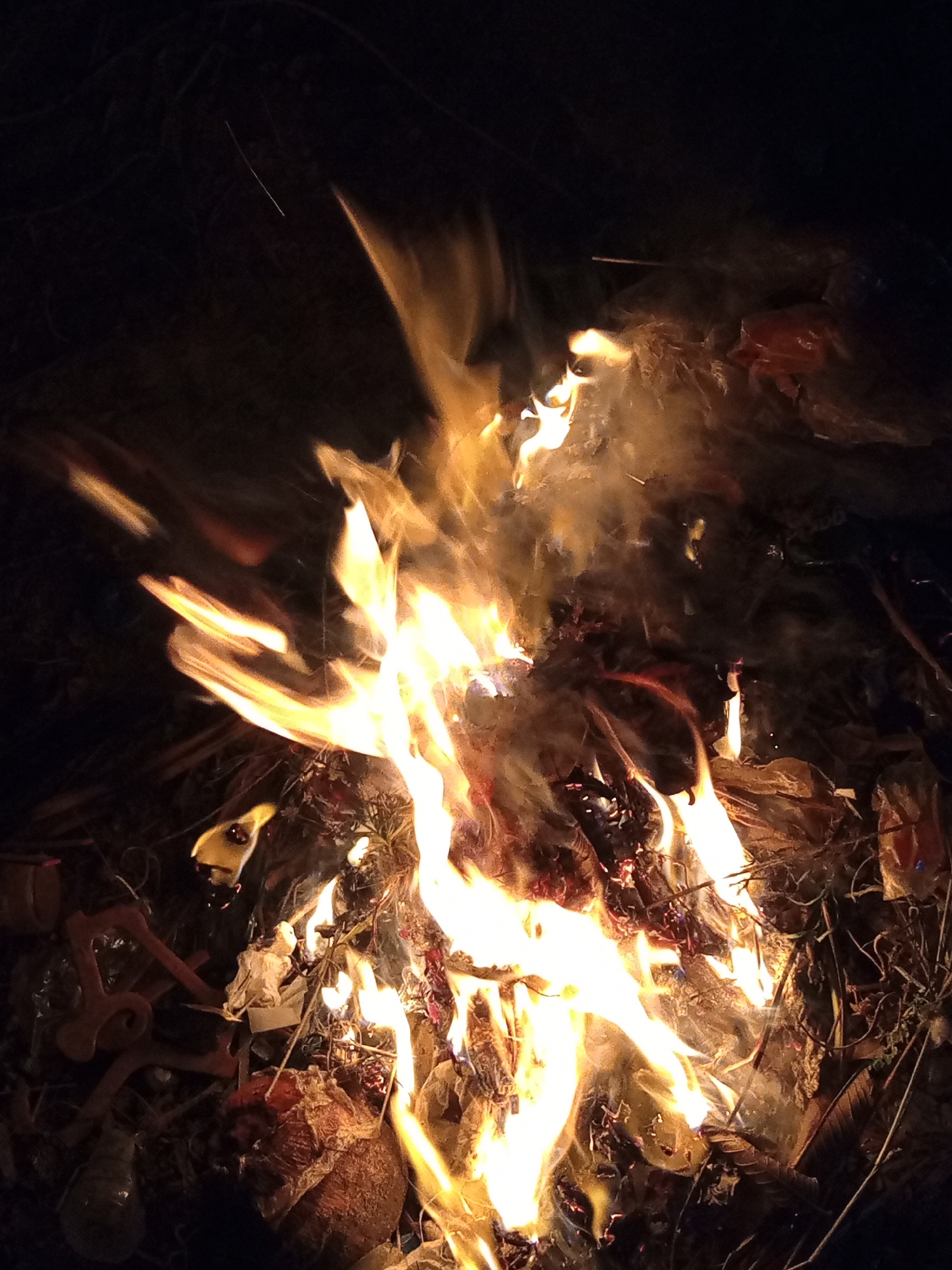 The image depicts a close-up view of a bonfire at night. The flames are bright yellow and orange, with some white at the hottest points, licking upwards. Smoke is visible, particularly in the upper right portion of the frame, tinged with orange by the firelight. The bonfire appears to be composed of dried vegetation, leaves, and possibly some discarded materials, as evidenced by the presence of what looks like plastic and possibly fabric in the lower left and central areas. A reddish-brown, textured object, resembling a dried fruit or root, is prominent in the lower center. The background is dark, suggesting a rural or outdoor setting, possibly a backyard or open space in Buketteukuh, Indonesia, given the context. There are no people visible in the image. The overall impression is one of a controlled burning of debris in a dark environment.