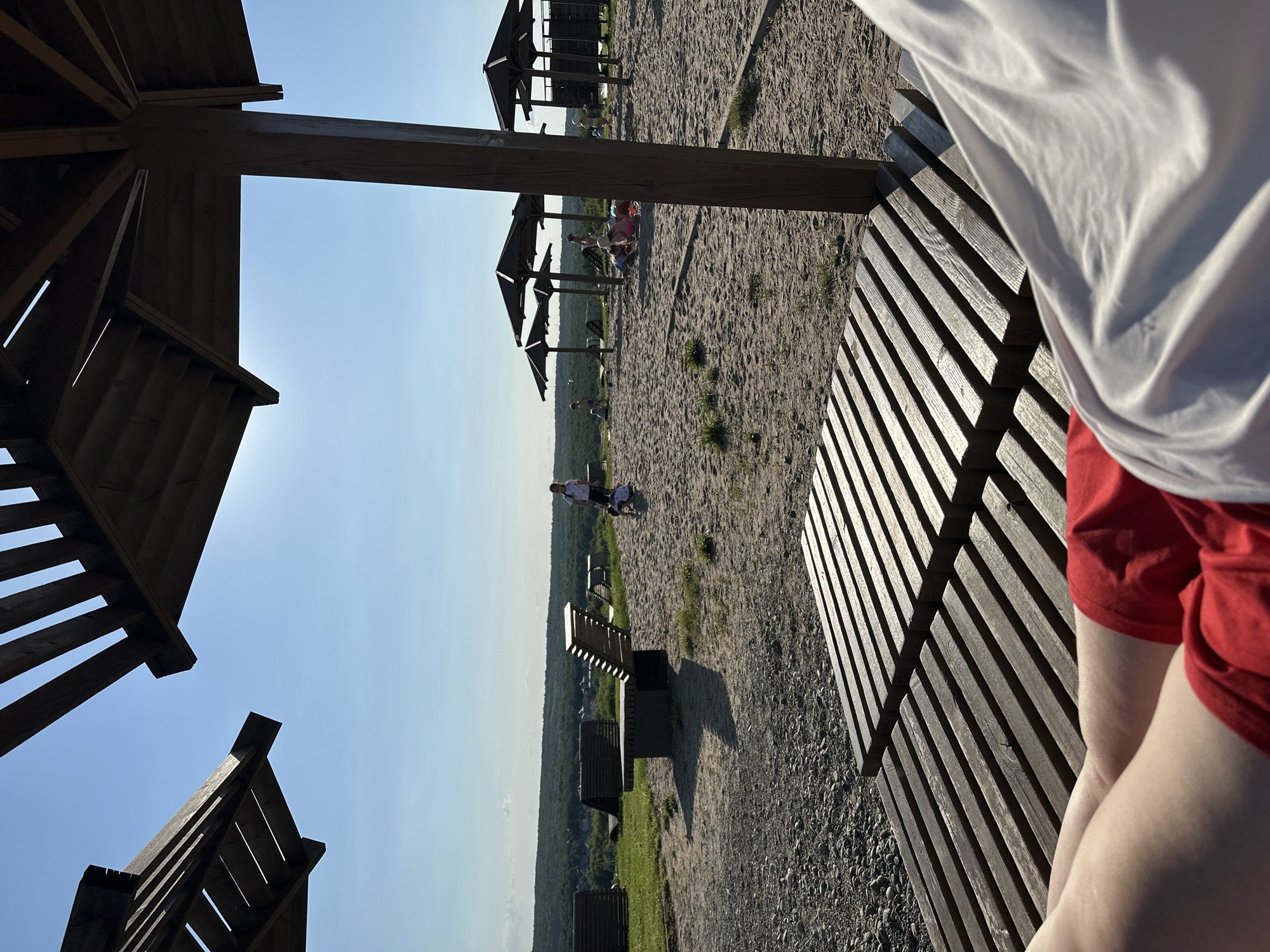 The image depicts a sunny day scene at a beach or sand-covered recreational area, likely located in Samara, Russia. In the foreground, a person's legs, clad in red shorts and partially covered by a white garment, are visible, resting on a wooden slatted surface. The wooden structure appears to be part of a bench or lounge chair.
Beyond the foreground, the sandy area is dotted with several wooden beach umbrellas, providing shade. In the middle ground, several people, including children, are engaged in various activities. Some children are playing in the sand, while others appear to be walking or standing. In the background, a panoramic view shows a lush green landscape with trees and a distant village or settlement, under a clear blue sky with the sun shining brightly. The overall impression is one of a relaxed, outdoor leisure environment.