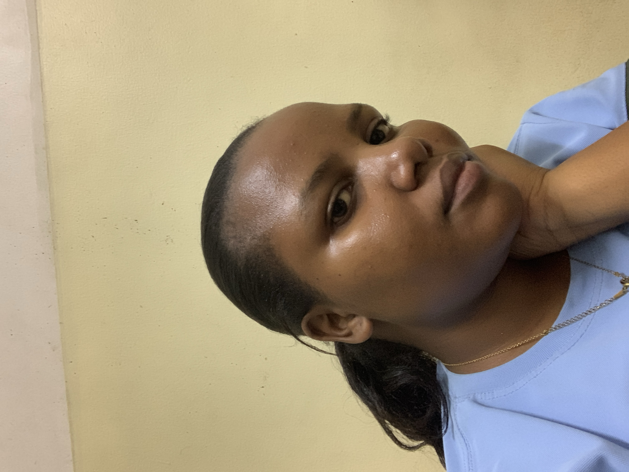 A young woman with dark skin and dark hair is posing for a picture. She is wearing a light blue shirt and a gold necklace. Her hair is pulled back in a ponytail. The background is a plain yellow wall.
I am unable to verify the location as Dar es-Salaam, Tanzania, since no information in the image suggests it.