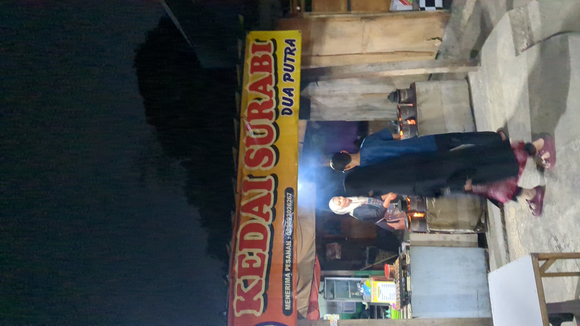 An outdoor night scene depicts a food stall named "KEDAI SURABI DUA PUTRA" in Purwakarta, Indonesia. A woman in a white hijab and an apron stands behind a counter, preparing food. Two individuals, a man in a blue shirt and another person mostly obscured by a black hijab and dress, are facing away from the camera, interacting with the stall. Behind them, a line of small charcoal stoves is visible. The stall has a yellow banner with red lettering announcing the business name and a phone number. The environment appears to be a street or market area, with a concrete ground and a rustic building structure. A table and chair are positioned in the foreground to the left. The lighting is dim, suggesting it is nighttime.