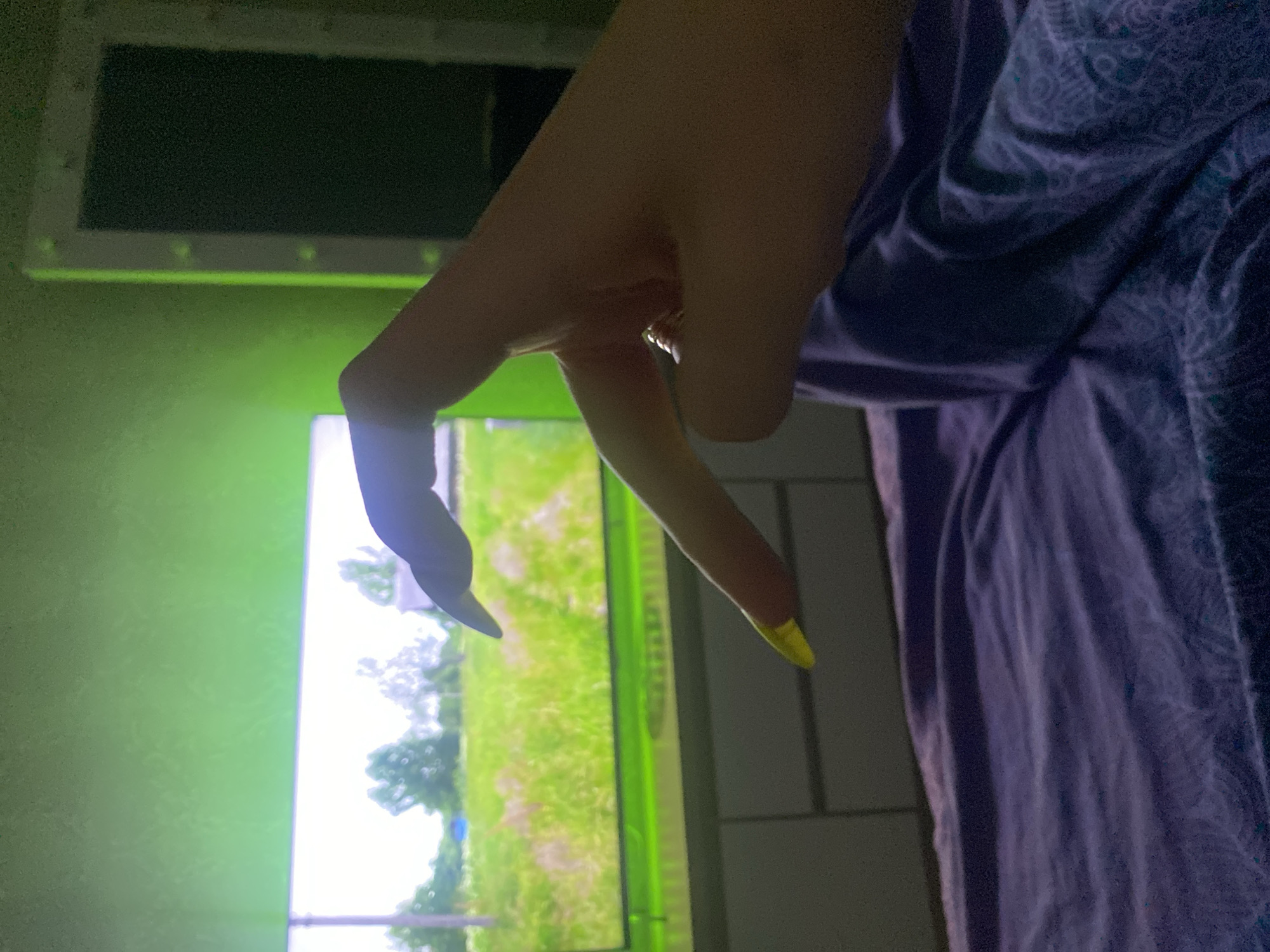 A hand with long fingernails painted black and yellow forms a peace sign against a background that appears to be a television screen displaying a grassy outdoor scene, possibly a park or field. The background also includes a tiled wall. The location cannot be determined to be the city of Chaikovsky, Russia from the image alone. The presence of patterned fabric suggests the hand is resting on a bed or sofa.