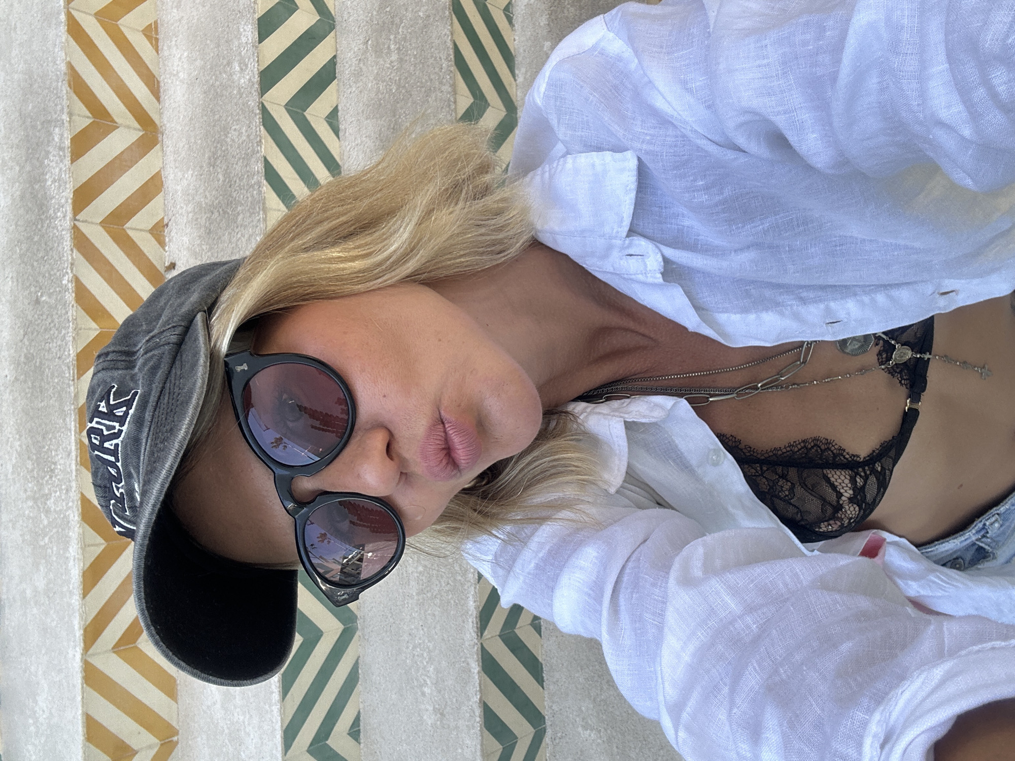 A woman with blonde hair and sunglasses is taking a selfie. She is wearing a white linen shirt open to reveal a black lace bralette and a necklace. She is also wearing a black baseball cap. The background is a patterned tiled staircase. The scene is set in North Kuta, Indonesia.