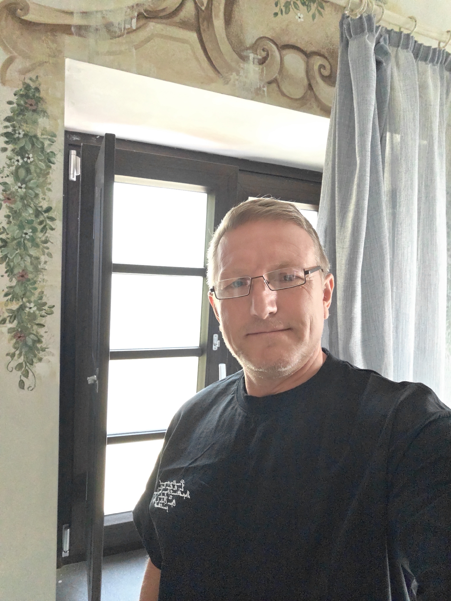 A man with short, blond hair and glasses is looking at the camera in a selfie. He is wearing a black t-shirt with white writing on the left chest. The background shows a window with sheer grey curtains on the right and a partially open dark wood door on the left. There is a fresco on the wall above the window and a decorative green vine with pink flowers painted on the wall to the left of the window. The lighting suggests it is daytime. The overall scene appears to be indoors, possibly in a residential or historical building. The man's facial expression is neutral.