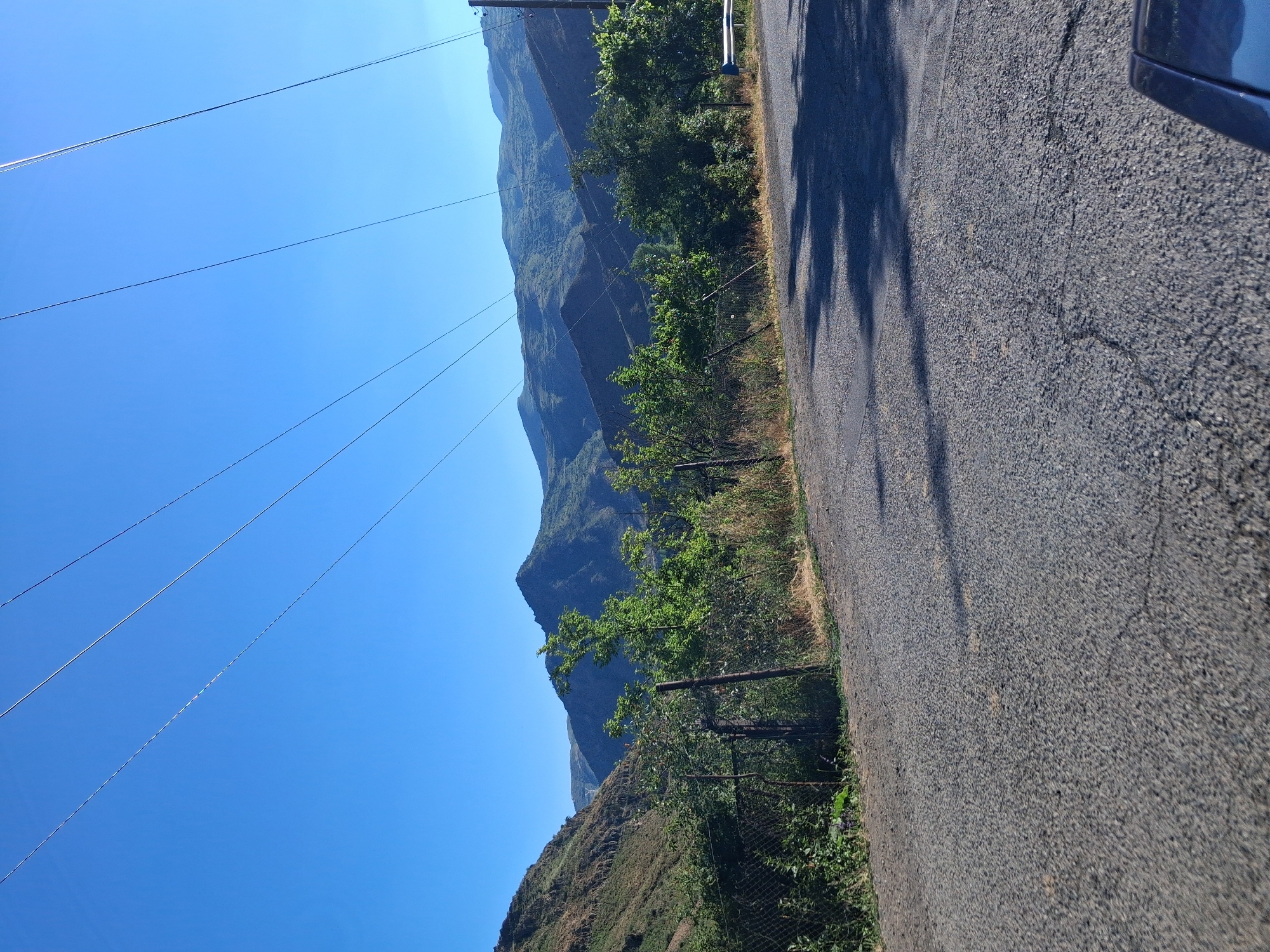 This image depicts a scenic mountain landscape in Shatin, Armenia. The foreground features a paved road with shadows cast by trees, suggesting it is daytime. Power lines crisscross the bright blue sky, indicating the presence of infrastructure in this mountainous region. To the left of the road, a fence separates the road from a vegetated hillside. Further back, rolling green mountains rise, some with visible terraces, hinting at agricultural activity or past human intervention. Lush green trees dot the landscape, especially near the fence line. The clear sky and bright sunlight suggest fair weather, likely mid-morning or afternoon given the shadow lengths. The overall impression is one of rural beauty and tranquility, typical of mountainous terrain in the region. There is no visible text in the image.