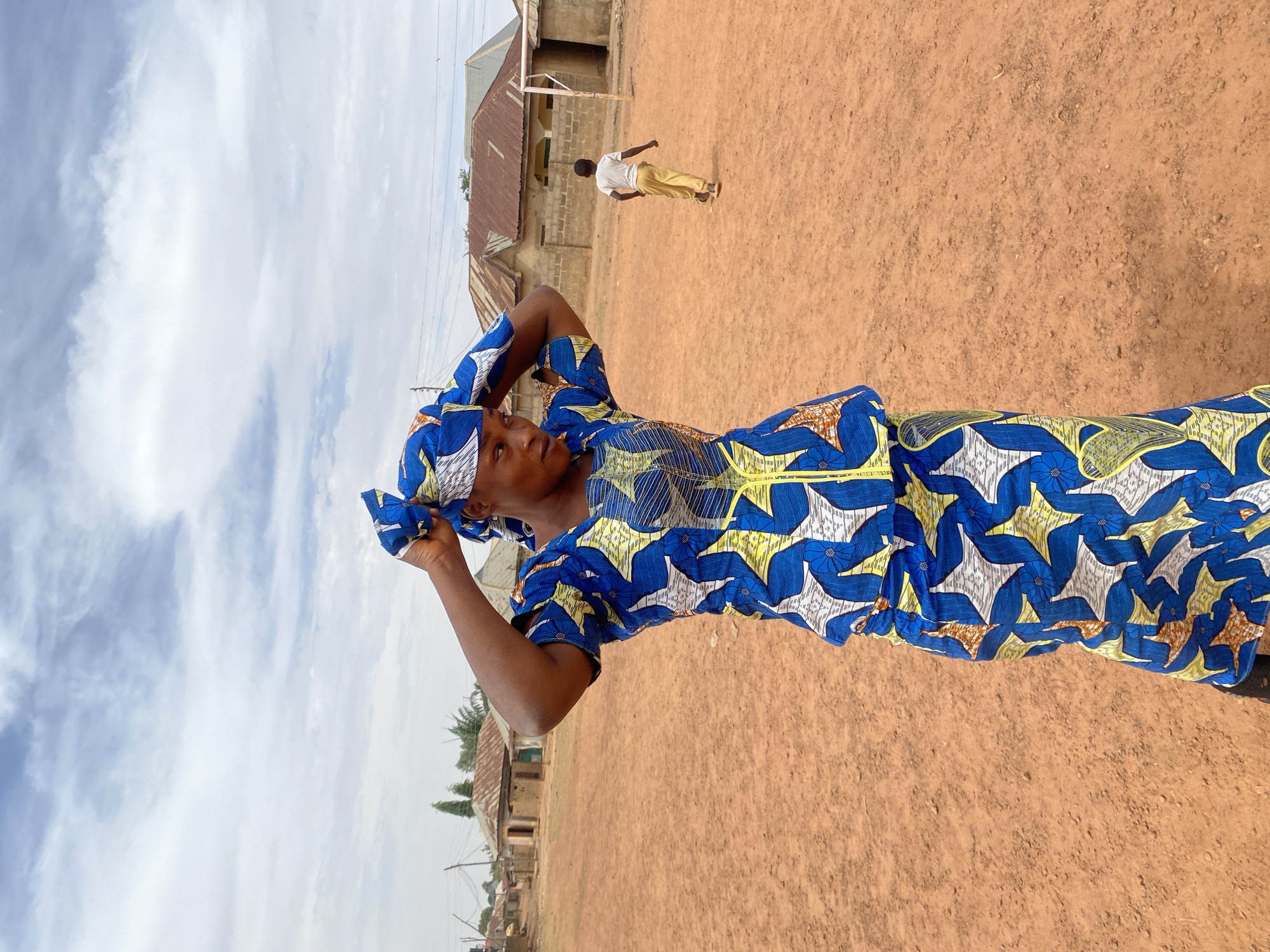 An outdoor scene captured during the daytime shows a person in traditional attire in the foreground, with an expansive unpaved area and simple buildings in the background under a cloudy sky.
The main subject, positioned on the right side of the frame (when upright), is an individual wearing a vibrant, patterned outfit. The attire features a striking design with dominant blue floral shapes, yellow wave-like patterns, and some white elements, consistent across a long dress or gown and a matching head covering. The person's skin tone suggests a person of color. Their arms are raised, and their hands are touching or adjusting the head covering, suggesting they might be tying it or arranging it. The person's gaze is directed slightly upwards and to their left.
The ground beneath is a reddish-brown, dry earth, characteristic of a dusty or unpaved area. In the mid-ground, a sparse landscape unfolds with several low-rise buildings constructed with what appear to be concrete blocks and topped with corrugated metal roofs. One building in the distance has a simple white goalpost in front of it, implying the area might be used as a makeshift playing field or community gathering space. Another person, seen from behind, is walking away from the camera towards these structures, dressed in a white t-shirt and light-colored pants.
The sky above is bright and shows a mix of white, scattered clouds against a lighter blue backdrop, indicating it is daytime. Some distant utility poles and power lines are faintly visible, along with sparse green foliage near the buildings. The overall impression is a vibrant, everyday scene in what appears to be a rural or semi-urban setting, likely in a warm climate. No text is visible in the image.