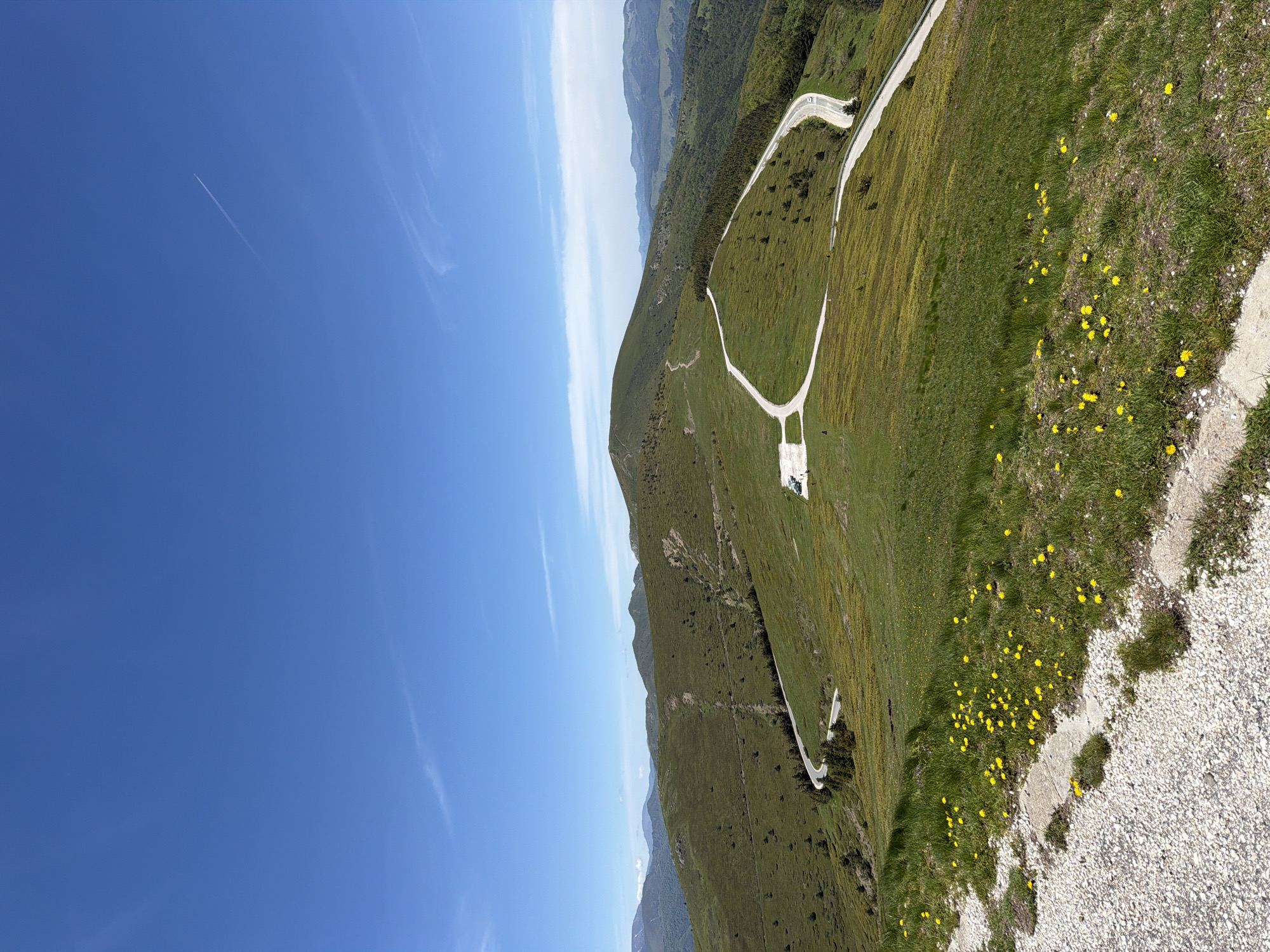 The image depicts a mountainous landscape under a clear, blue sky with wispy contrails. A narrow road winds across a green hillside dotted with yellow wildflowers. The location is identified as Beli Osam, Bulgaria. The environment consists of green hills and open sky. There are no people apparent in the picture, only the landscape and the path winding through it.
