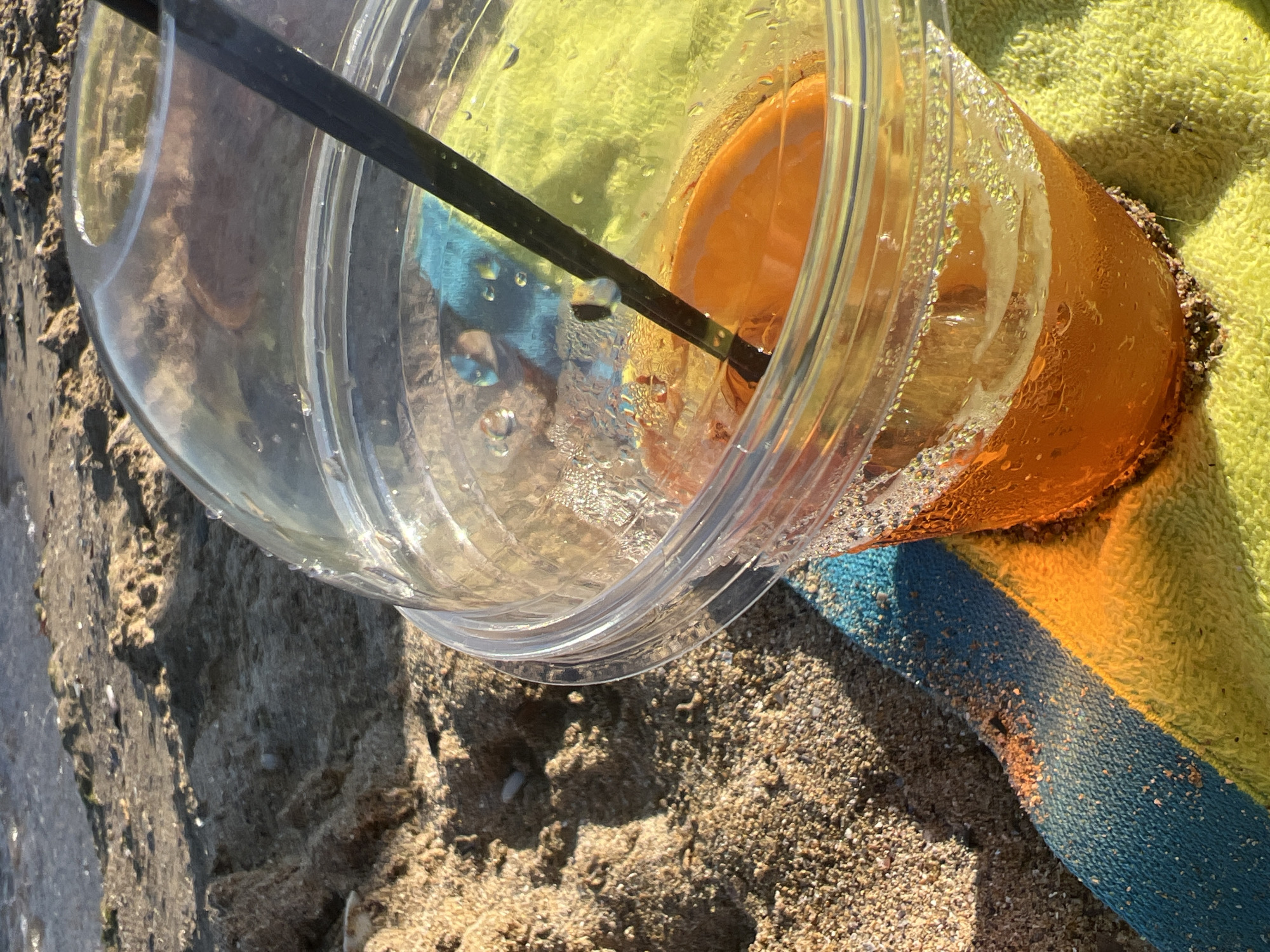 A plastic cup filled with an orange beverage and an orange slice sits on a striped towel on a sandy beach. A black straw is in the cup. The ocean is visible in the background. This image was taken in Fontanka, Ukraine.