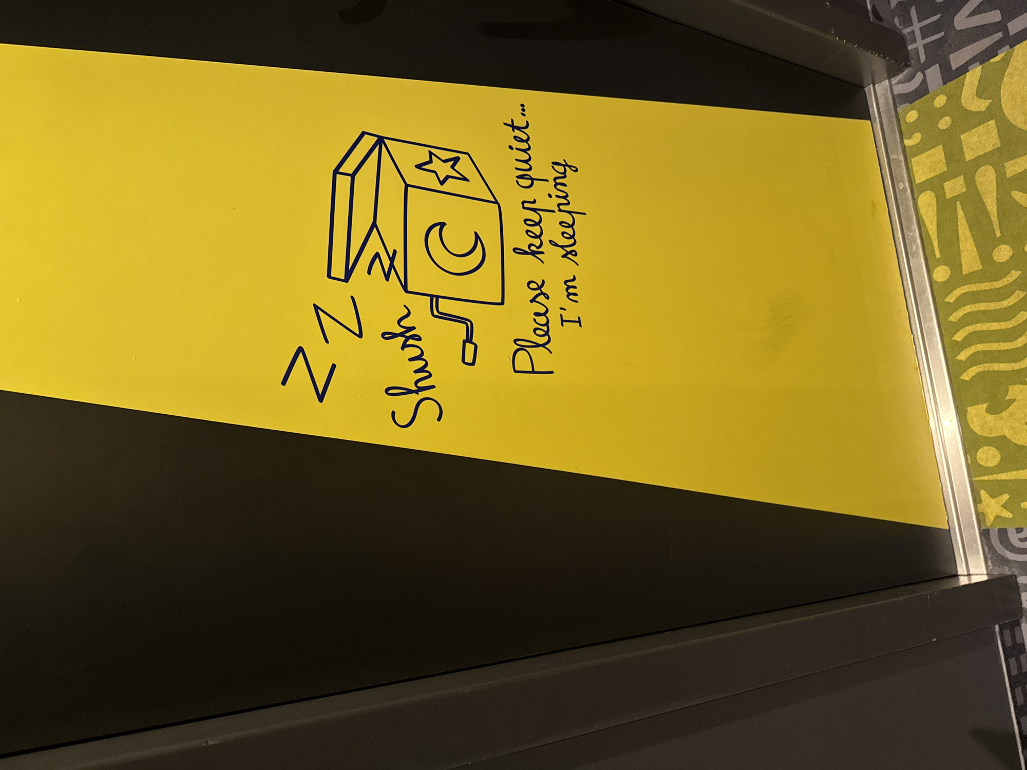 The image shows a close-up, eye-level shot of a dark grey elevator door with a bright yellow panel on the right side. The yellow panel features a blue ink drawing of a jack-in-the-box with its lid open, a crescent moon, and a star. Above the drawing, there are three "Z"s, indicating sleep, and the word "Shush" is written below them. Further down, the phrase "Please keep quiet... I'm sleeping" is written in a cursive font. The bottom of the image shows the floor, which has a grey and black patterned carpet on the left and a bright green mat with yellow abstract shapes and patterns on the right, near the elevator. The elevator is likely located in Stein, Netherlands, based on the context provided.