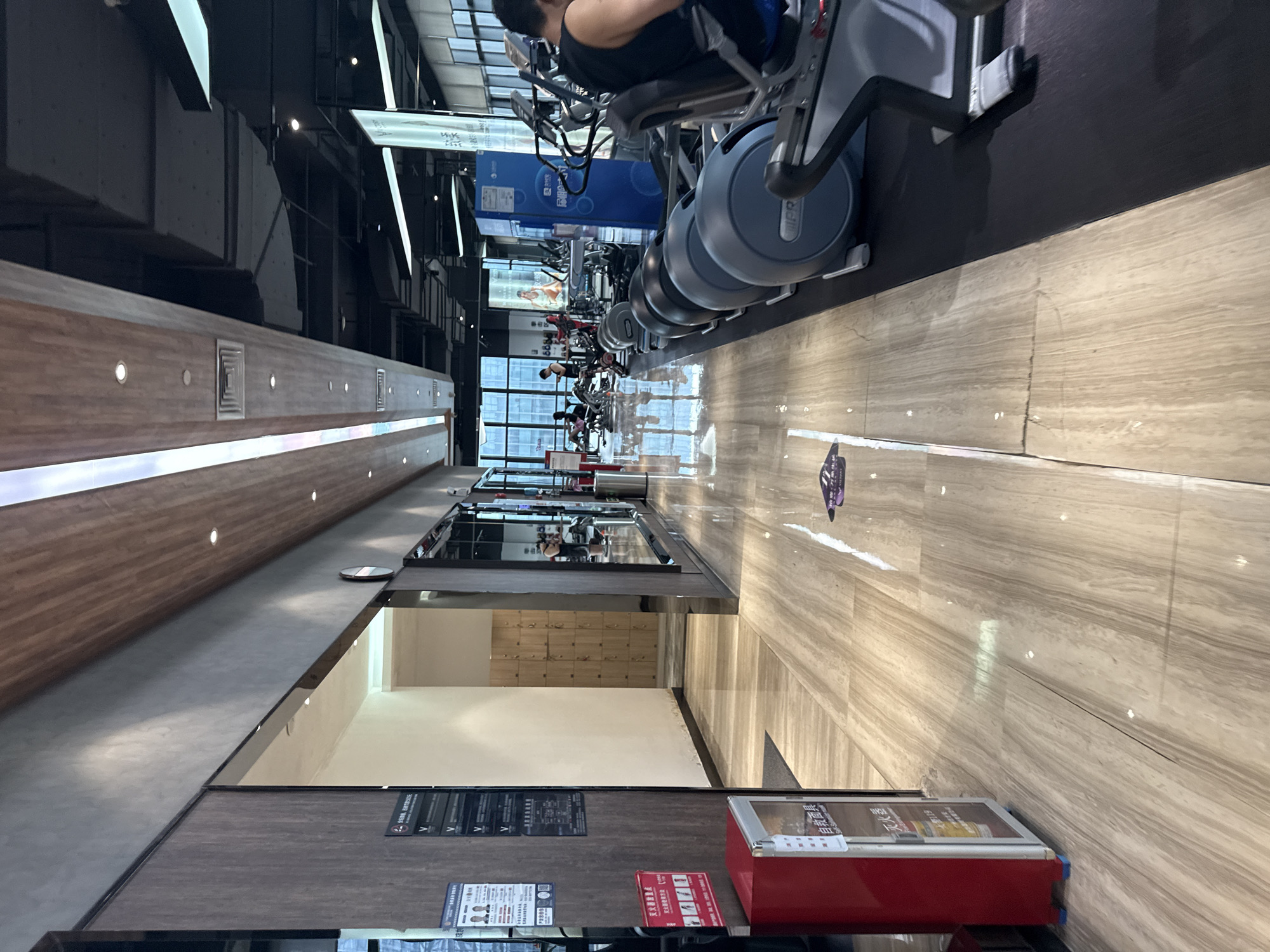 The image depicts the interior of a gym in Haizhu District, China. The gym features polished wooden floors that reflect light, leading into a section with black rubber matting where exercise equipment is located. Along the left side, there is a wall with wooden paneling and a glass-fronted display case containing a red dispenser. A hallway with lockers is visible further down the left side. On the right, a row of elliptical exercise machines is arranged, with a person seated on one of them. The ceiling is high, with exposed beams and lighting fixtures. Natural light enters through large windows in the background, revealing office buildings outside. Several people are seen in the background, engaged in various activities within the gym.