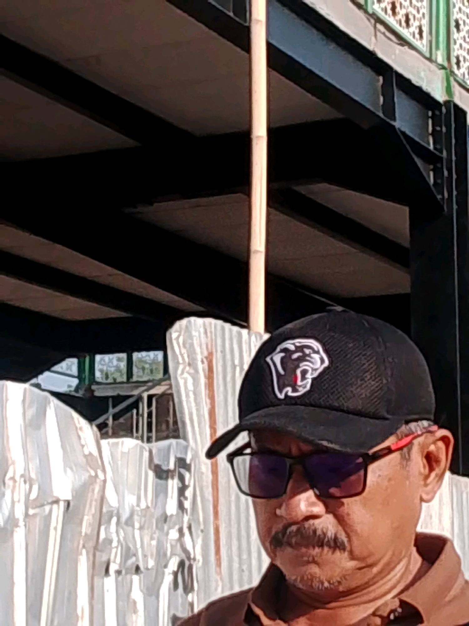 A man wearing a black baseball cap with a white bulldog logo and red-rimmed sunglasses is shown from the chest up. He has a mustache and is looking down. He is standing in front of white corrugated panels. In the background, there is a dark metal structure with visible beams, possibly an overpass or a building under construction. A thin bamboo pole is visible to the left of the man. The scene appears to be outdoors in a city environment. The lighting suggests it is daytime.