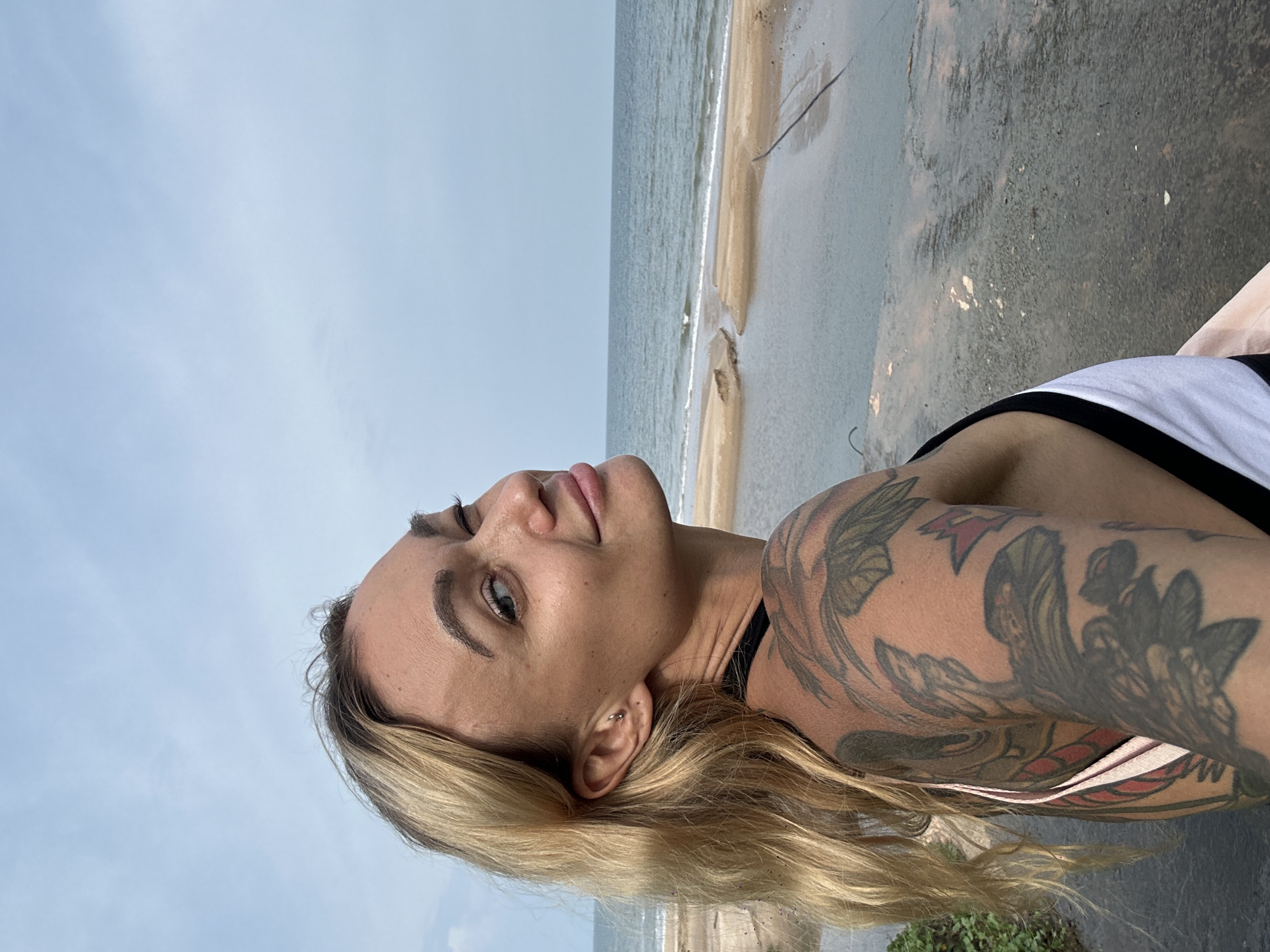 The image is a portrait of a woman, oriented 90 degrees clockwise, taken outdoors at a beach location. The main subject is a woman with light skin and long, flowing blonde hair, looking upwards and slightly to her left with a soft, content expression and a subtle smile. She has well-defined dark eyebrows, and appears to be wearing light makeup, including eyeliner and lip gloss. A small piercing is visible in her left ear. She is wearing a top with black and white sections. Her left arm and shoulder are prominently featured, covered in intricate, colorful tattoos depicting what appear to be birds and floral or nature-inspired motifs.
The background depicts a tranquil beach scene. On the left side of the image, a vast, light blue-grey sky, indicative of an overcast or hazy day, fills much of the frame. On the right side, the calm, light blue-grey sea meets a sandy shore with gentle waves. Beyond the immediate beach line, there is a darker, possibly damp or rocky section of ground, with some minor debris or vegetation visible. The lighting is soft and diffused. The location is Tien Thanh Ward, Vietnam. There is no visible text in the image.