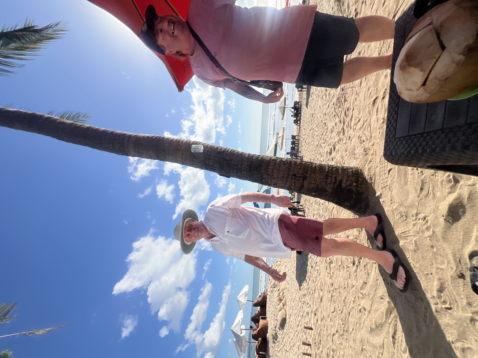 Two men are on a sandy beach in Kuta, Indonesia, under a clear blue sky with scattered white clouds. The man on the left, wearing a white polo shirt, maroon shorts, a light-colored fedora, and black flip-flops, walks towards the camera. The man on the right, wearing a pink t-shirt, dark shorts, and a dark baseball cap, smiles and looks towards the camera. He has a dark strap across his chest and tattoos on his arm. A palm tree is visible between the two men, and a red umbrella is positioned above the man on the right. In the background, beach chairs, umbrellas, and lounge chairs are set up, and the ocean is visible with a few people in the water. The sand has footprints and debris. In the foreground, a portion of a black woven table is visible, with a halved coconut on top.