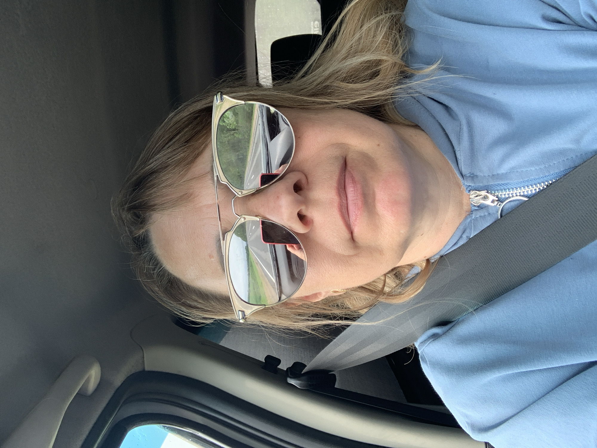 A woman is sitting in the driver's seat of a car, wearing sunglasses and a light blue hoodie. The sunglasses reflect a road stretching into the distance with trees on either side. A red phone is also visible in the reflection. The woman's hair is blonde and shoulder-length. The car appears to be on a road with a seatbelt fastened across her chest. The scene is outdoors during the daytime. The location is estimated to be in the vicinity of Хаценчыцкі сельскі Савет, Belarus.