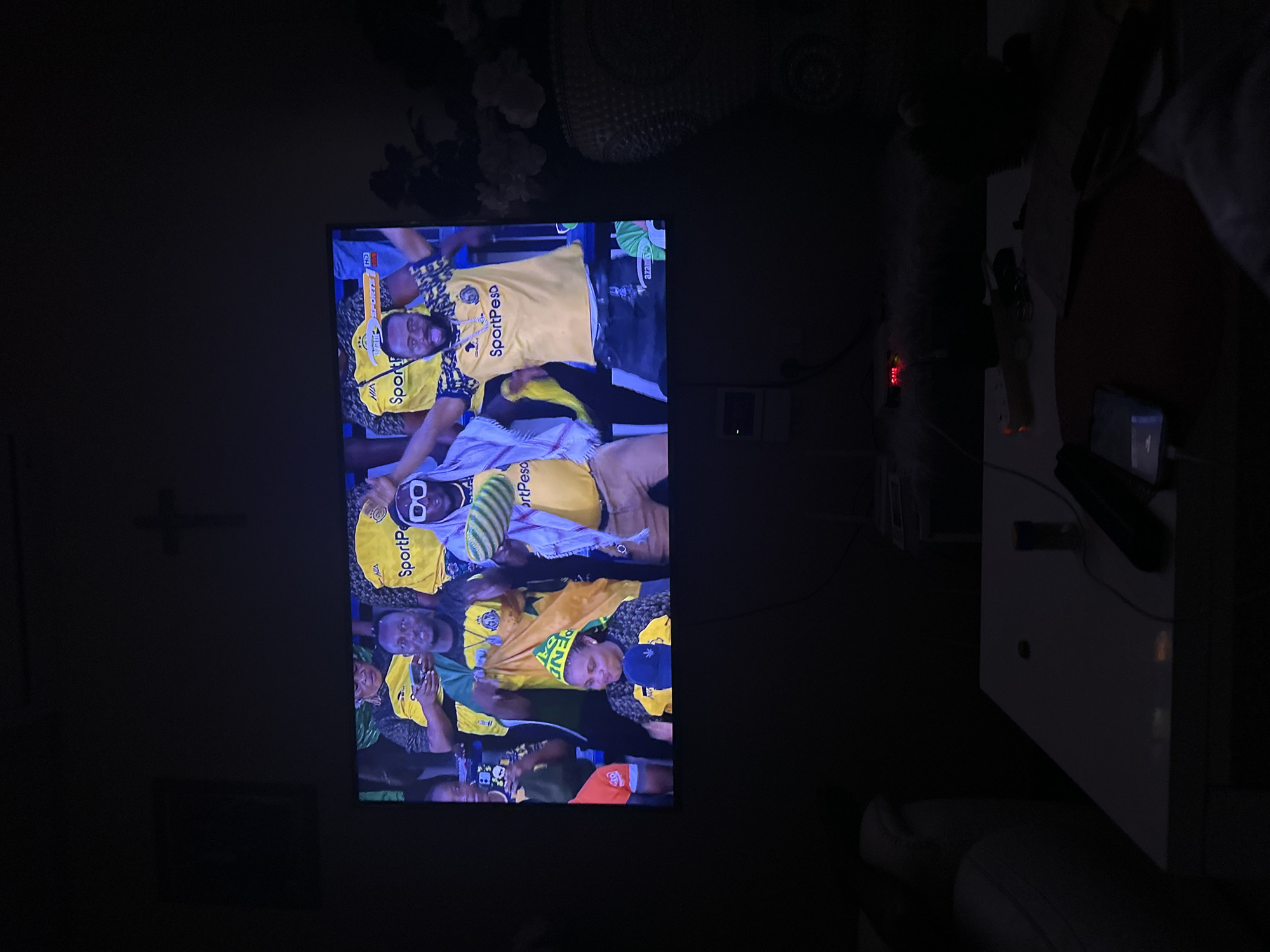 The image depicts a television screen displaying a live sports broadcast, likely a football match, in a dimly lit room. On the screen, several individuals, predominantly male, are visible wearing yellow and black sports jerseys with the "SportPesa" logo. One person in the foreground is holding a striped object, possibly a towel or scarf, and another is wearing distinctive white-rimmed glasses. The individuals appear to be fans or players celebrating or engaged in the game. The screen also shows logos for "SportPesa" and "HD." The surrounding environment is dark, with a hint of a white couch on the left and a table with some objects on the right, including a phone and what appears to be a remote control. A small digital clock on a shelf displays "04:15." A crucifix is subtly visible on the wall above the television. While the specific location in Dar es Salaam, Tanzania is not explicitly stated, the presence of "SportPesa," a prominent betting company in East Africa, and the context of a sports broadcast strongly suggest a setting within Tanzania.