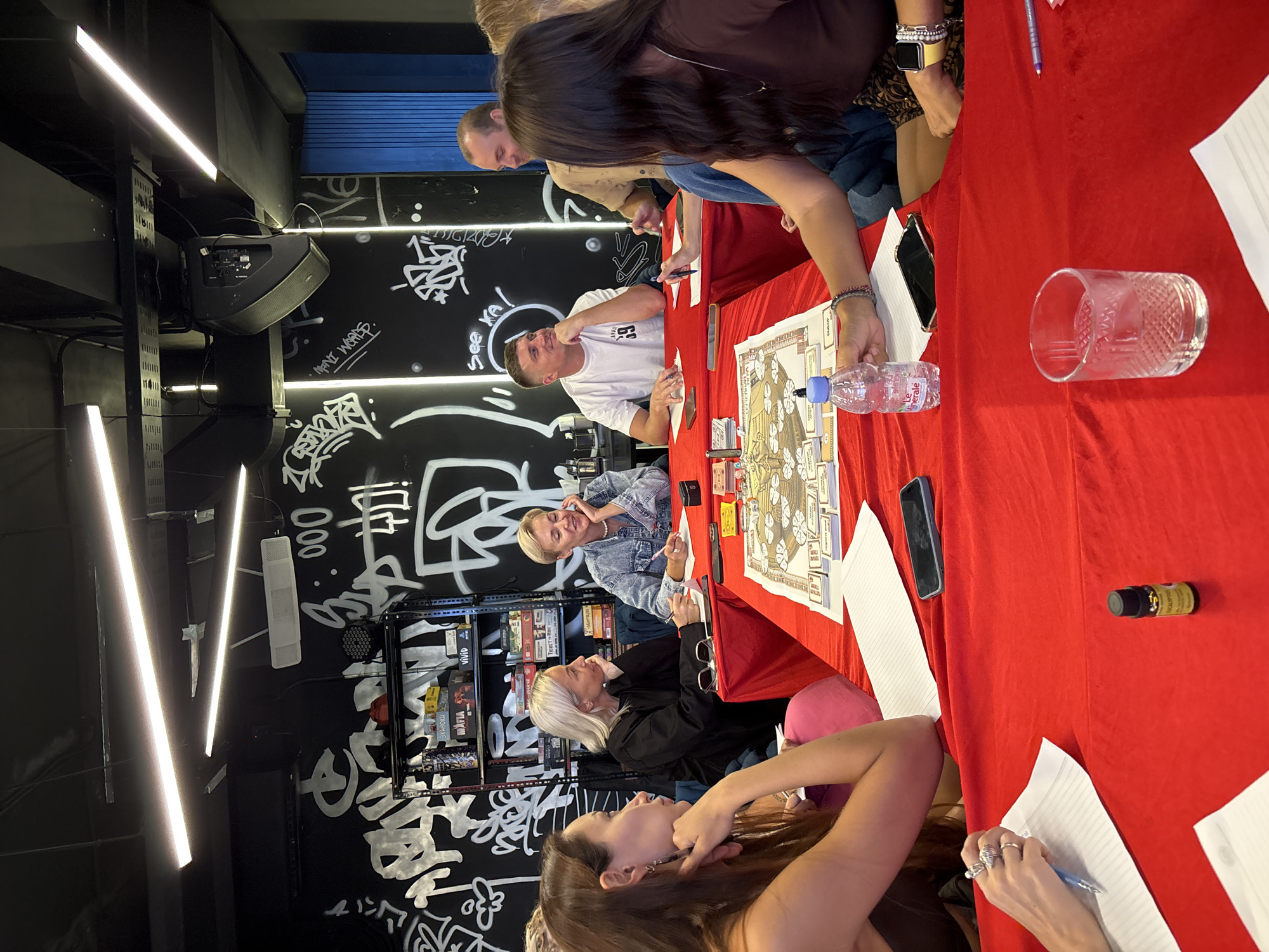 The image shows a group of approximately 8 people sitting around a long table covered in a red cloth, likely indoors, possibly in Kuta Selatan, Indonesia. Some of them are engaged in what appears to be a board game or activity involving paper and writing implements. A board game is visible on the table, along with water bottles, glasses, phones, and other small objects. The background features a black wall with white graffiti, shelves containing various items including what appear to be board games, and a sound system. Above, there are long, linear lights. The scene suggests a casual, social gathering focused on the board game activity.