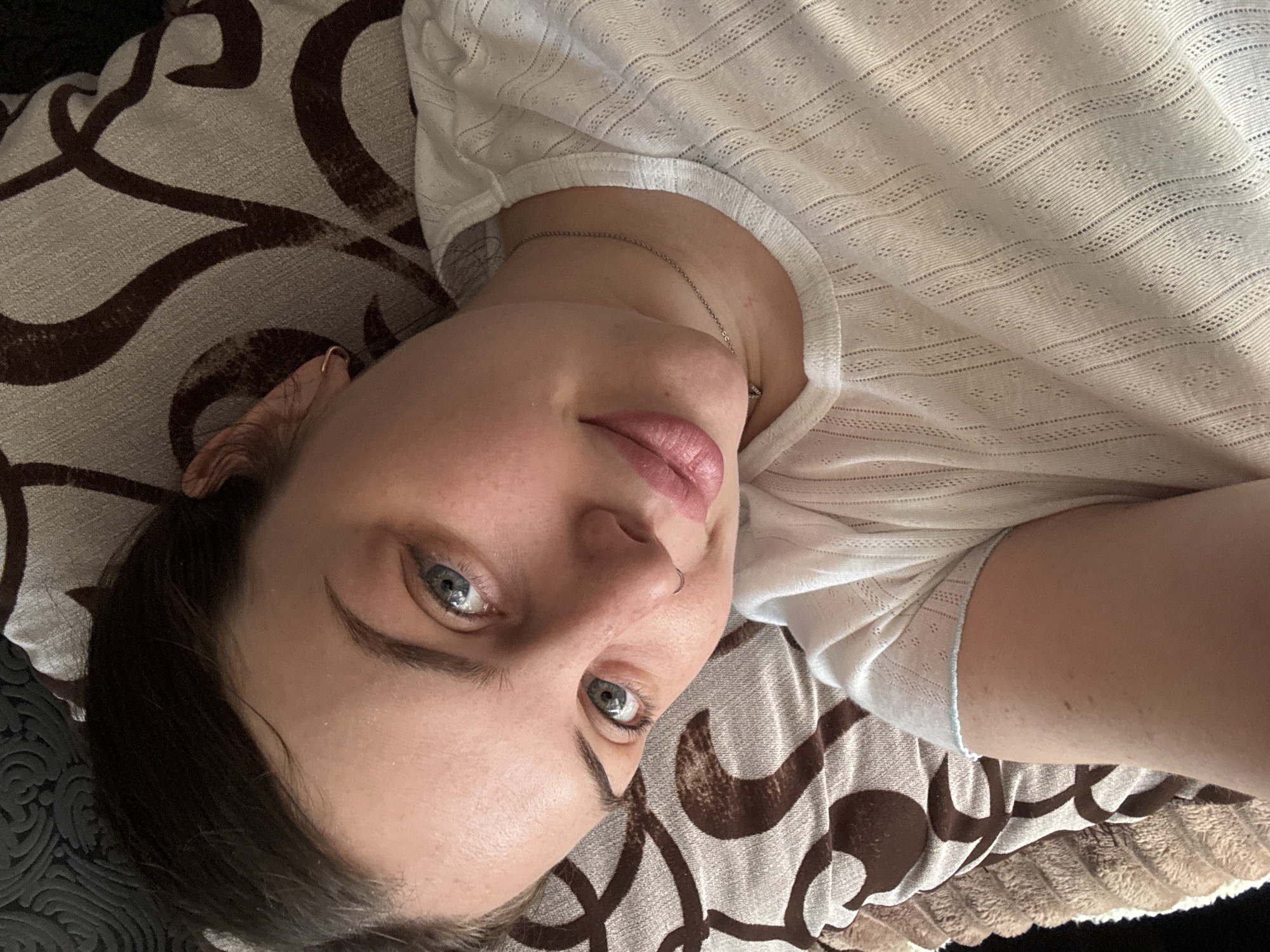A young woman is lying on a patterned cushion or pillow, possibly a couch or bed. She is wearing a white, lacy or textured short-sleeved shirt. She has fair skin, dark hair, and light-colored eyes. A thin silver chain necklace is visible on her neck. The background is a brown and tan patterned fabric. The image does not contain enough information to locate the location of the photo in Chornomorskoe, Ukraine.