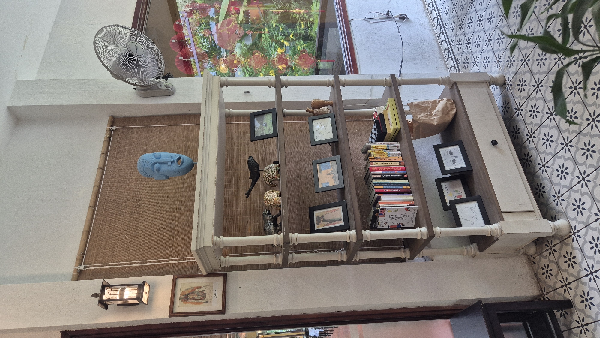 The image depicts an indoor scene in Đà Nẵng, Vietnam, likely a cafe or a retail space. A tall, multi-tiered shelf unit with a distressed white and wood finish stands against a white wall. The shelves are adorned with various decorative items and books.
On the top shelf, a blue mask-like sculpture is prominently displayed above a bamboo blind. To its left, a framed artwork hangs on the wall, illuminated by a wall-mounted lantern. The second shelf from the top holds a collection of small decorative objects, including what appear to be elephant figurines and a bird sculpture, alongside framed photographs. The third shelf also features framed pictures. The bottom shelf is filled with an assortment of books, with spines of various colors and sizes. On the right side of the bottom shelf, a brown paper bag is casually placed. A drawer is integrated into the base of the shelf unit, with a single dark knob.
The environment is characterized by the tiled floor with a repeating floral pattern in shades of grey, black, and white. A large window to the right offers a view of an outdoor area with lush greenery and red floral decorations hanging outside, suggesting a festive or vibrant atmosphere. A white oscillating fan is mounted on the wall above the window. A dark wooden stool is visible to the left of the shelf unit. The overall impression is one of curated display and a well-decorated interior space.