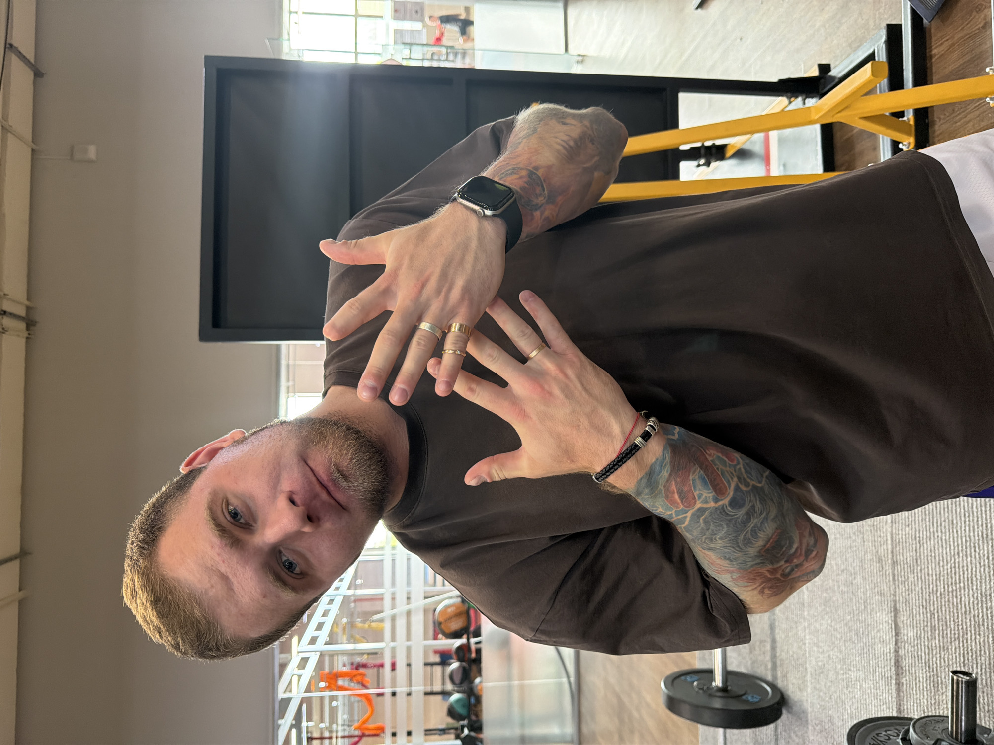 A man with light brown hair and a beard, wearing a dark brown t-shirt, stands in a gym. He is leaning to his left, looking at the camera with a playful expression. His left arm is bent upwards, with his fingers extended, revealing several rings on his fingers and a black braided bracelet on his wrist. A tattoo covers his left arm and extends up to his shoulder. His right arm is also bent upwards, with his hand open. He is wearing a smartwatch on his left wrist. In the background, a black panel stands on a yellow metal frame, and weightlifting equipment, including a barbell plate and dumbbells, are visible on a grey carpeted floor. The gym appears to be in the city of Chaikovsky, Russia, as indicated by the context of the request.