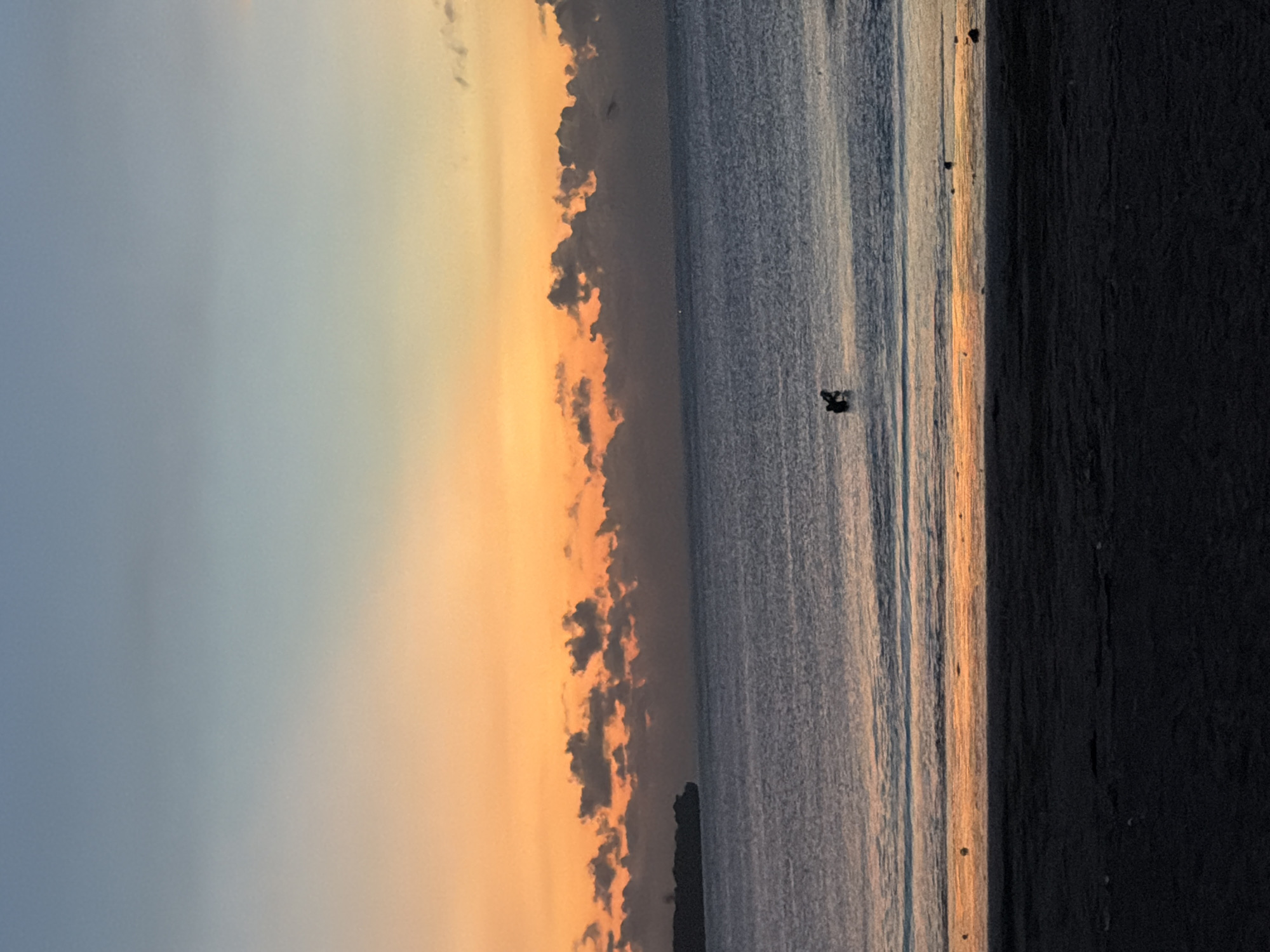The image captures a serene beach scene, likely at sunrise or sunset, in Jimbaran, Indonesia. The sky transitions from a soft blue to a warm orange hue, with scattered dark clouds along the horizon. The ocean reflects the colors of the sky, creating a shimmering effect. There are a few small figures visible in the water, possibly people. The beach itself is dark, possibly due to the early morning light or the color of the sand. The scene suggests a tranquil atmosphere.