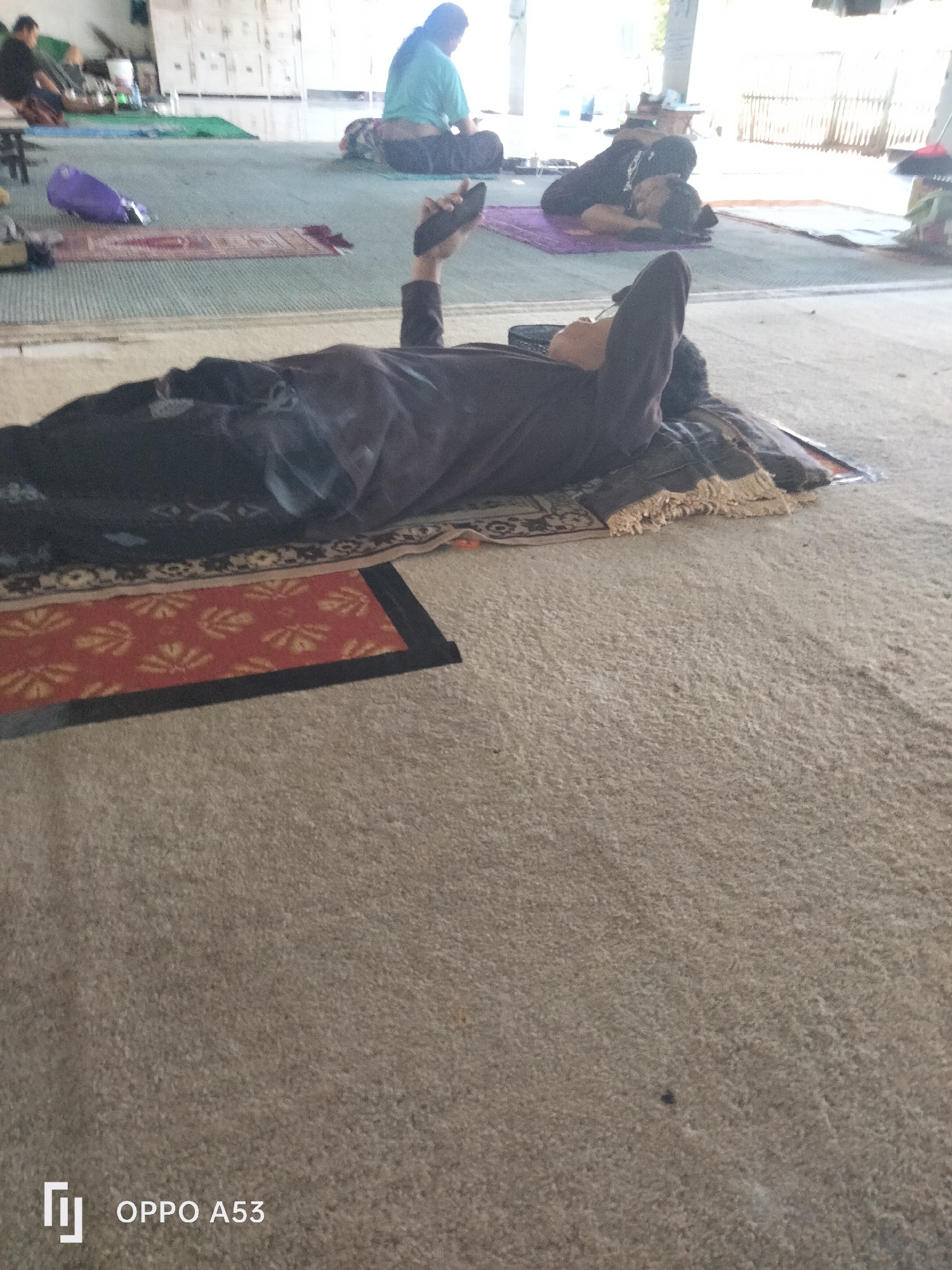 In an indoor area resembling a mosque or prayer hall in Badung, Indonesia, an individual is seen lying on their back on a prayer mat. The person is wearing dark clothing and holding a black object, possibly a phone, in their right hand above their head. The floor is covered with a light-colored carpet. In the background, other individuals are visible in various states of rest or prayer, some wearing head coverings. The environment appears to be a communal space with natural light filtering in from the right side, illuminating the scene. There are also prayer mats and other prayer-related items scattered around. The overall impression is of a peaceful and communal gathering for prayer or rest.