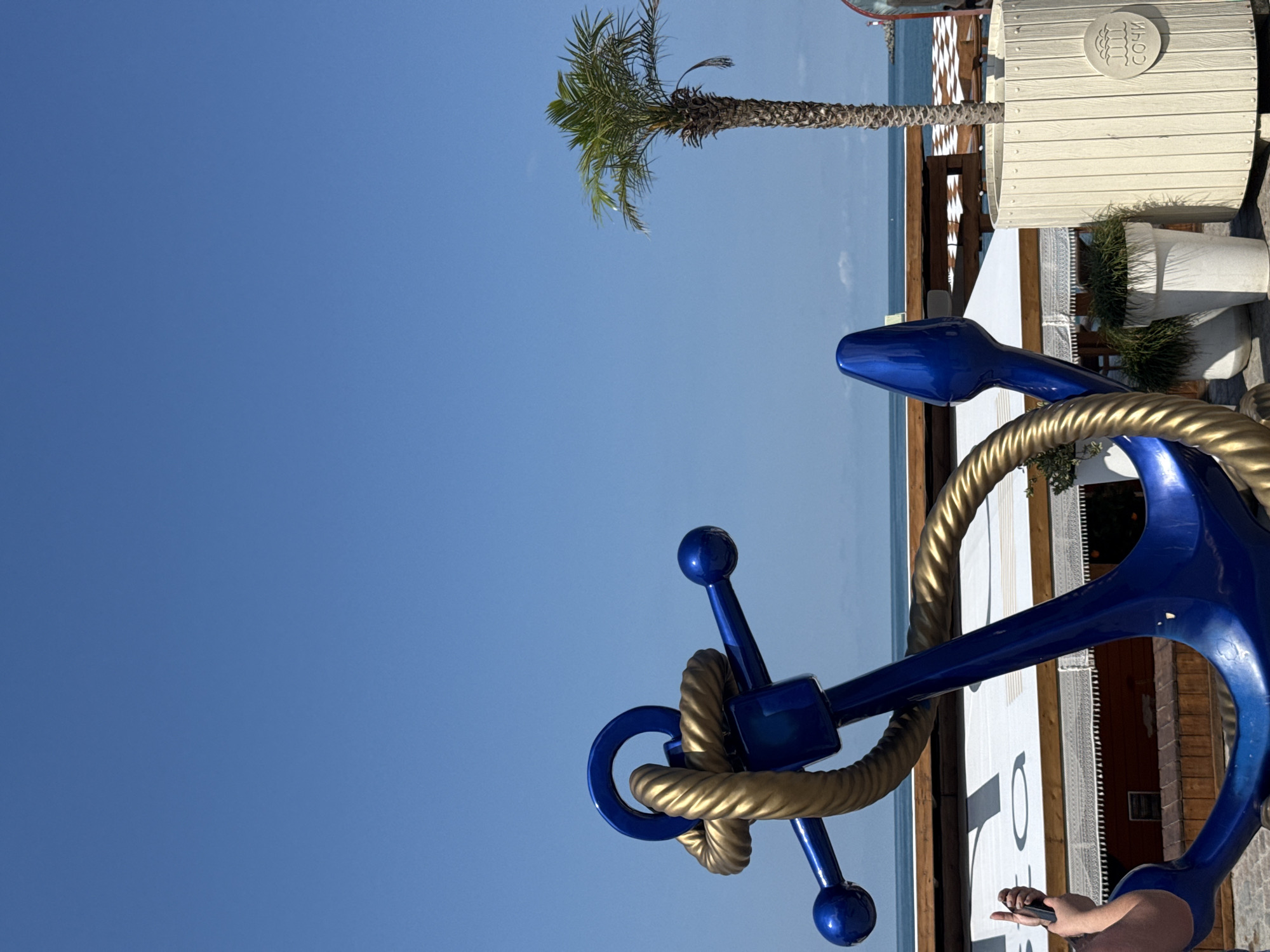 This image depicts a close-up of a large, bright blue anchor sculpture with a golden rope coiled around it, set against a clear blue sky. The anchor is positioned in the foreground and appears to be a decorative element, possibly at a seaside resort. In the mid-ground, a palm tree stands tall, to the right of the anchor. Behind the palm tree and the anchor, there is a glimpse of the sea and a beach umbrella or canopy. The background reveals a wooden structure, possibly part of a beach cafe or seating area. A white planter box with the word "СОЧИ" (Sochi) embossed on it is visible next to the palm tree, indicating the location as Sochi, Russia. A person's hand holding a black smartphone is partially visible in the lower left corner of the frame, suggesting someone is taking a photograph of the scene. The overall scene is bright and sunny, characteristic of a summer day by the sea.