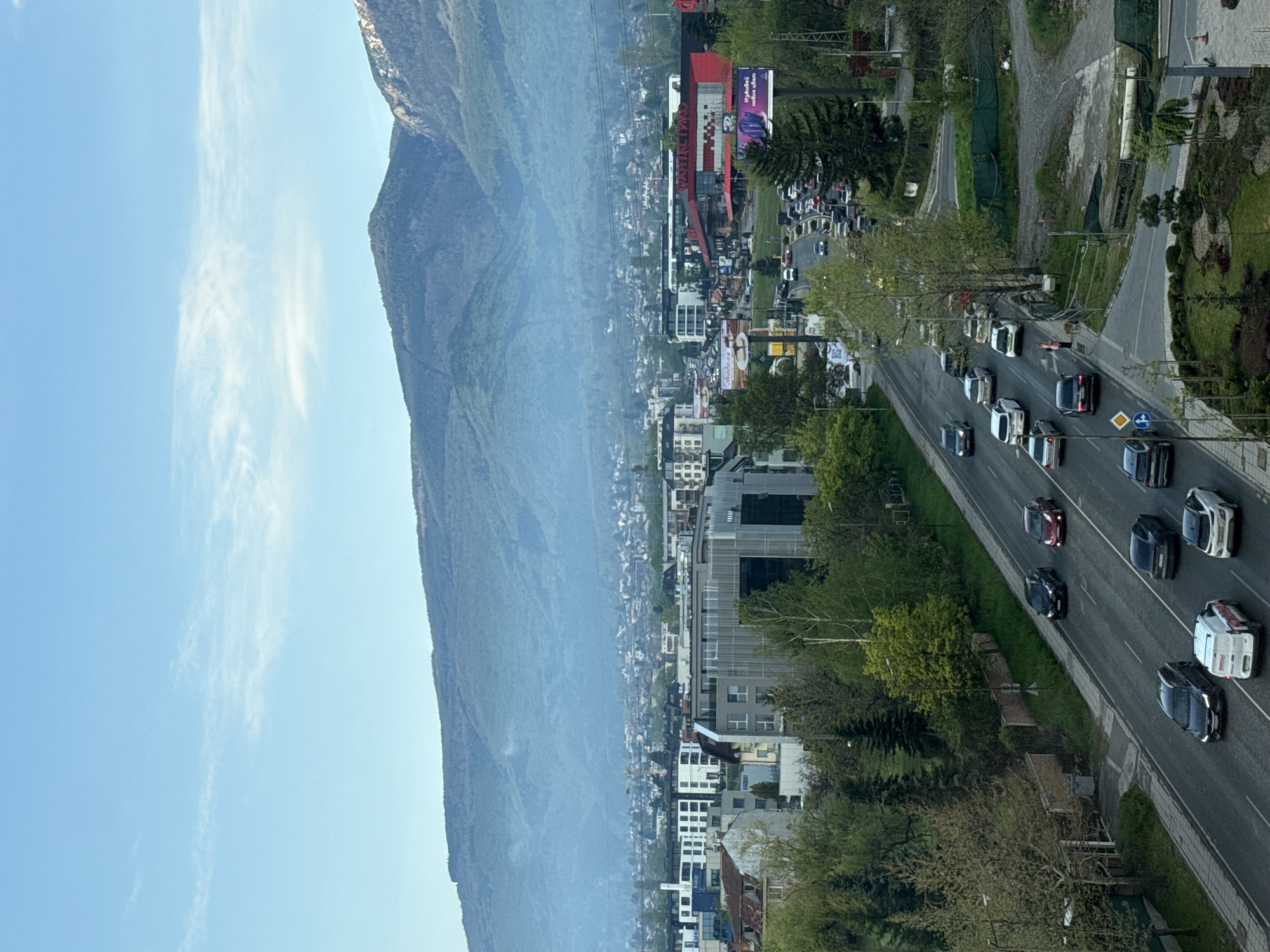 This image, captured from an elevated perspective during daytime, presents an urban landscape set against a majestic mountain backdrop. The scene is rotated 90 degrees clockwise and should be viewed upright.
In the foreground (right side when viewed upright), a multi-lane asphalt road carries moderate to heavy traffic, with numerous cars visible in both directions. Many vehicles appear to be stopped or moving slowly. Green verges and trees line the road, separating it from a paved sidewalk where a person in a pink top can be seen. Road signs, including a yellow diamond with a black right-pointing arrow and a blue circle with a white upward arrow, are visible on the median.
The middle ground is occupied by a dense urban area featuring a mix of commercial and residential buildings. A prominent red building with a large sign clearly displays the name "TECHNOMARKET." Another billboard nearby shows text in what appears to be Bulgarian, though it is not fully legible from this distance.
Dominating the background (left side when viewed upright) is a large, forested mountain range, its slopes covered in greenery and receding into a slight atmospheric haze. The sky above is clear and blue, dotted with a few thin, wispy white clouds. The bright, even lighting suggests it is late morning or early afternoon. The presence of a "TECHNOMARKET" sign supports the provided location context of Sofia, Bulgaria.