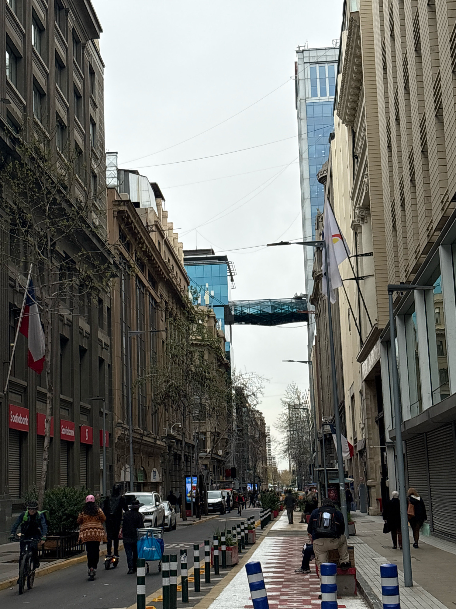 The image depicts a street scene in Santiago, Chile. On the left side of the street, a dark gray building with visible vertical columns and numerous windows dominates the foreground. A flag, white with a blue square in the upper left corner and a red stripe on the right, hangs from the building. Further down the street, two red signs with white text that reads "Scotiabank" are visible.
On the right side of the street, a lighter-colored building with a textured facade and ornate architectural details runs parallel to the left-side building. Two flags are mounted on this building; one is white with a symbol and the other is the French flag. Above these buildings, a modern glass skybridge connects two taller structures.
The street itself features a pedestrian walkway with a red and white striped pattern. Several people are present, including a person on a bicycle on the left, and others walking or riding scooters in the middle of the street. Cars are parked or driving on the right side of the street, which also has a sidewalk with more pedestrians. The sky is overcast.