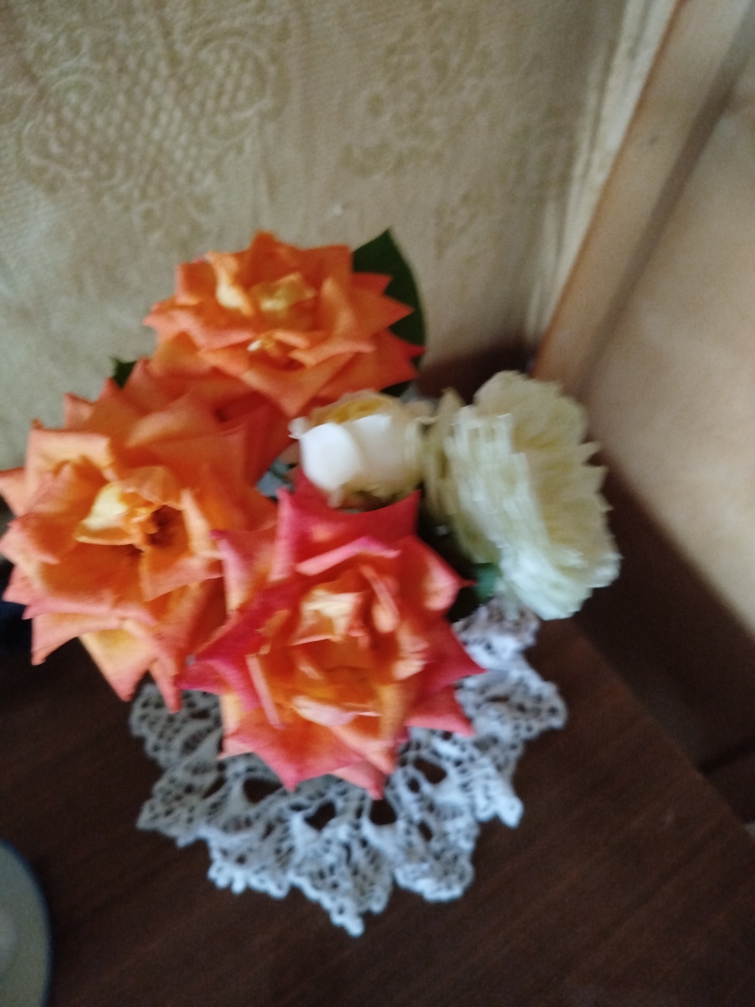 The image shows a close-up of a bouquet of roses on a lace doily placed on a wooden surface. There are several orange and pink roses, along with some white flowers. The background appears to be a textured wallpaper. The lighting is dim, casting soft shadows. The scene is indoors, likely on a table or shelf. There is no visible indication of the city of Romny, Ukraine.