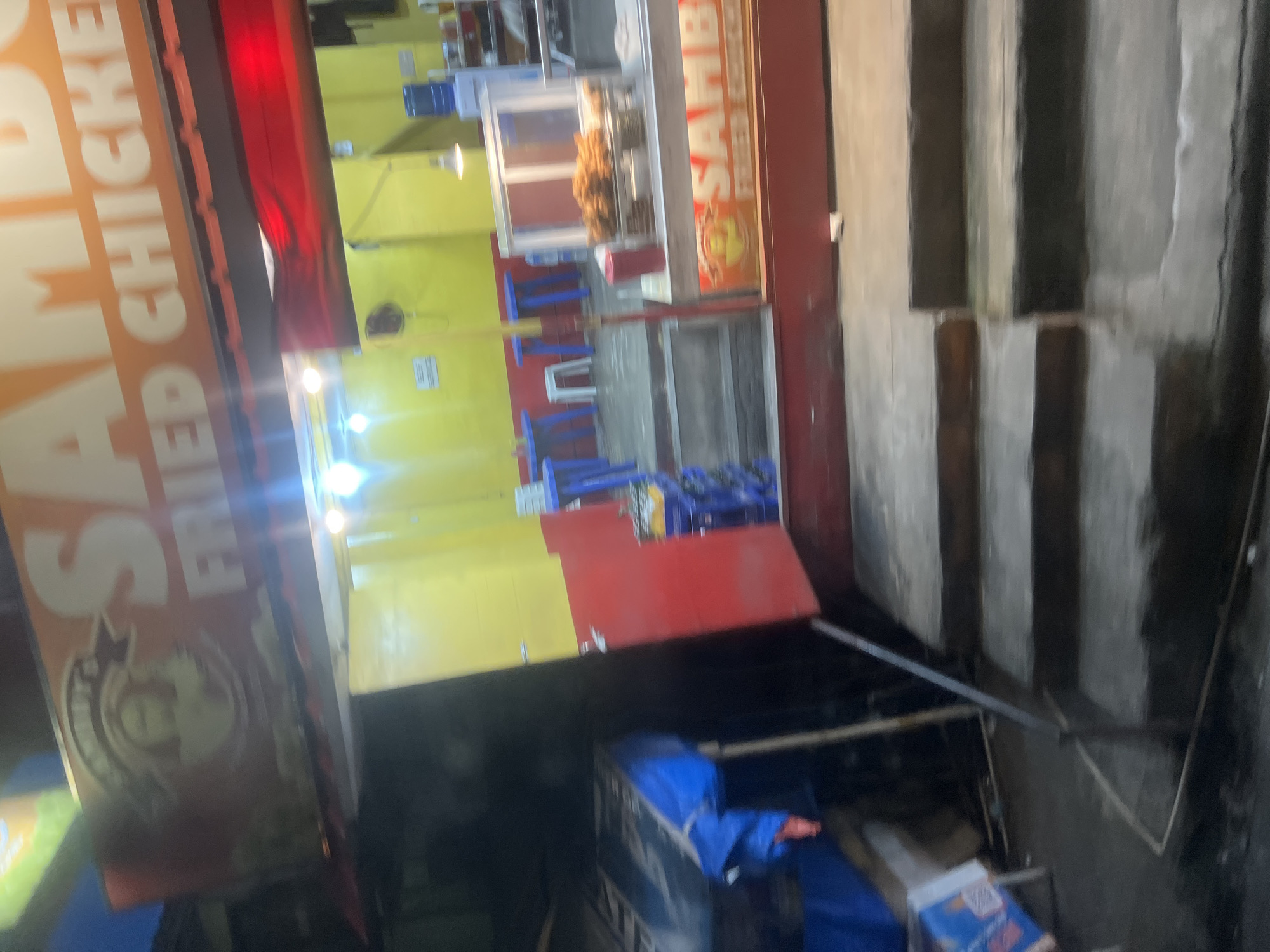 The image shows the exterior of a food establishment with prominent signage for "SAMBO'S FRIED CHICKEN." The establishment has a bright yellow facade with red accents. There are blue plastic chairs and tables set up, suggesting an outdoor seating area, though the overall scene appears to be dimly lit, possibly indicating evening or nighttime. A glass display case showcases fried chicken. In the foreground, a large sign with white lettering on an orange background reads "SAMBO'S FRIED CHICKEN." The image is slightly tilted, affecting the perspective. The setting appears to be a street or sidewalk in Davao City, Philippines, with a concrete staircase visible on the right side of the image. There's a blue tarp covering some items in the lower portion of the frame. The overall atmosphere is that of a casual, local eatery.