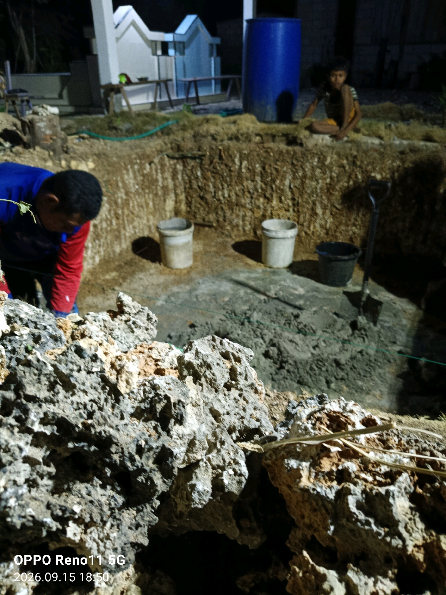 The image shows a construction site at night, possibly in the city of Otan, Indonesia. A person in a blue and red shirt is working near a dug-out area. There are two buckets and a shovel in the pit. In the background, a large blue barrel and a young boy are visible. The environment appears to be uneven and rocky, with some vegetation. A white structure resembling a tomb or monument is also visible in the background. The timestamp "OPPO Reno11 5G 2026.09.15 18:50" is present in the bottom left corner.