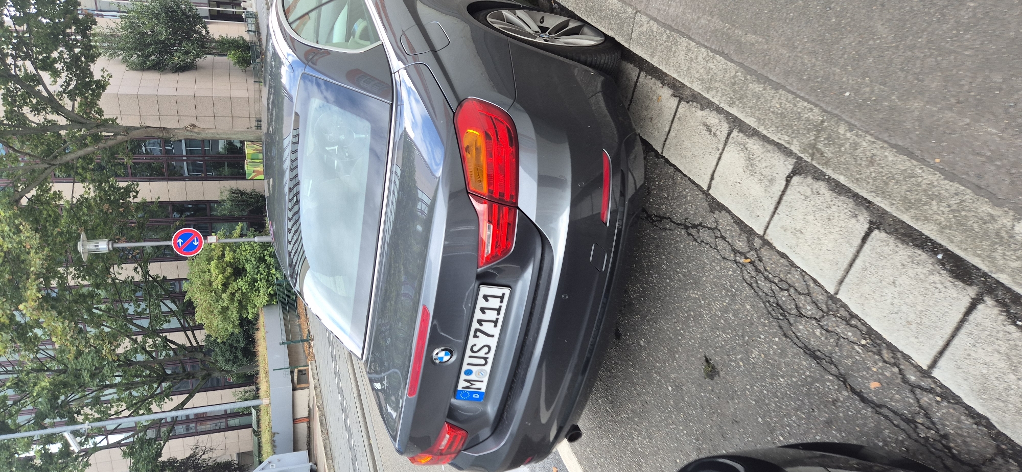 A dark gray convertible BMW is parked on the side of a street in Mainz, Germany. The car's license plate reads "M US 7111". In the background, a modern building with a glass facade and trees can be seen. There is also a street sign with a red circle and blue arrows indicating a no-passing zone. The street is paved, and there is a concrete curb and sidewalk. The car is parked at an angle to the curb. The weather appears to be overcast.