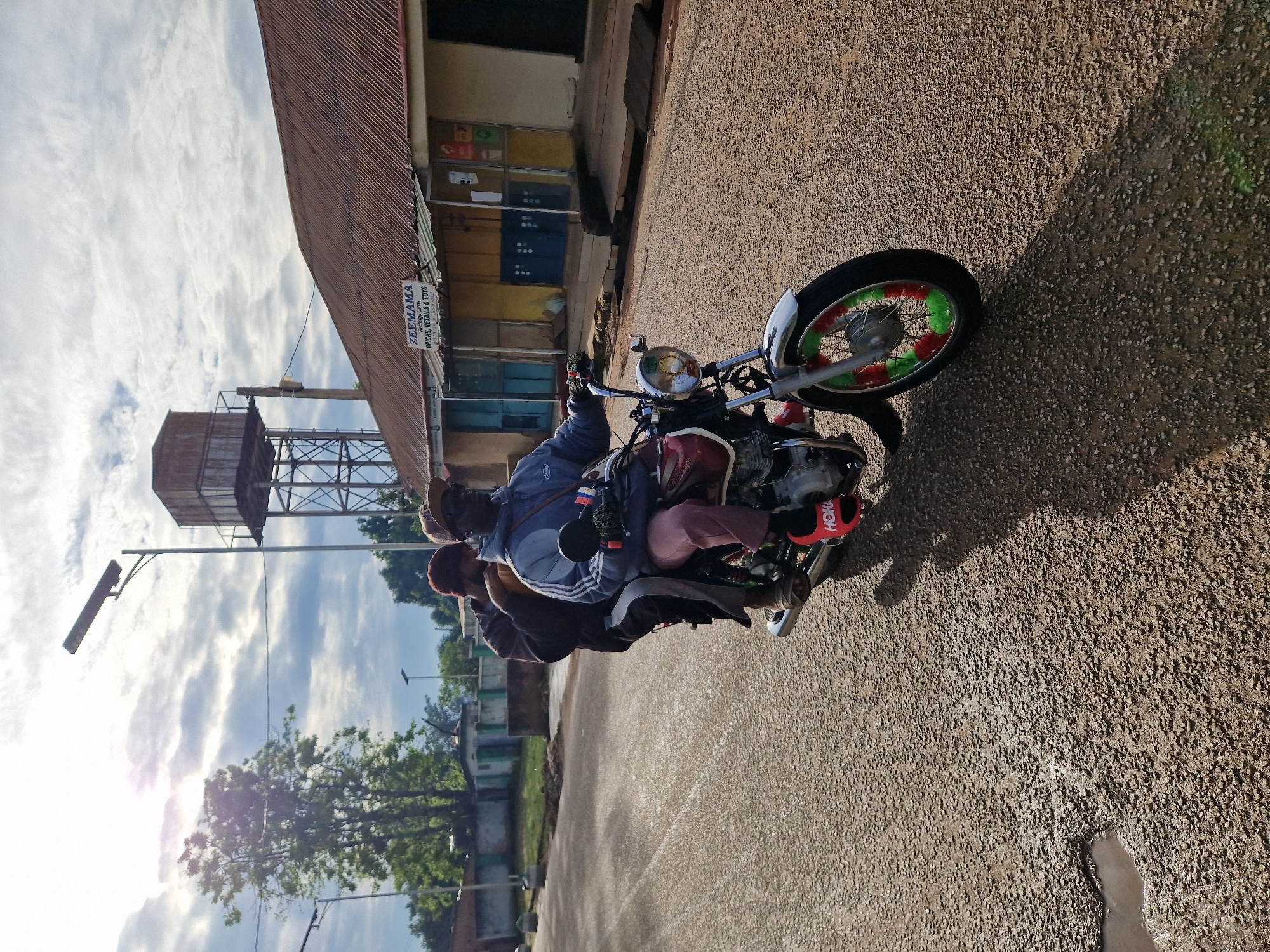 The image depicts two individuals on a motorcycle, traveling on a paved road in Kabong, Nigeria. The rider, wearing a blue jacket, a cap, and sunglasses, is at the front, while a passenger in a maroon beanie and dark jacket sits behind him. The motorcycle is red and silver, with green and red accents on the front wheel. The road is wet in places, with a puddle visible on the left. In the background, there are buildings with corrugated iron roofs, one of which has a sign that reads "ZEEMAMA Recharge Cards, Bricks, Retails & Toys". A water tower stands behind the buildings. The sky is partly cloudy, with the sun shining through, casting shadows. The overall scene suggests a typical day in a Nigerian town.