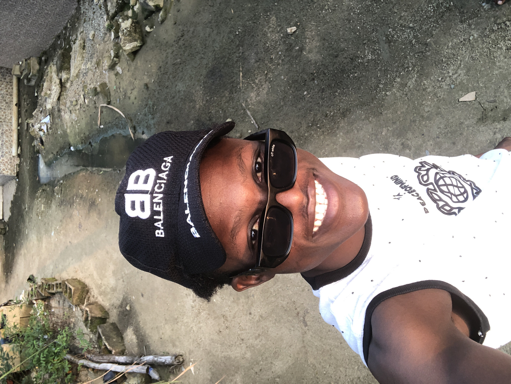 A young Black male is taking a selfie outdoors. He is wearing a black Balenciaga baseball cap, black sunglasses with "off white" written on the side, and a white sleeveless shirt with a black trim and a design on the front. He has dark skin and is smiling, showing his teeth. The background shows a dusty path with some overgrown vegetation and what appears to be a drainage ditch or stream running through it. The area looks somewhat unkempt, with scattered rocks and debris. The overall setting suggests an urban or semi-urban environment in a warm climate. This scene could be a street or alleyway in Ado, Nigeria.