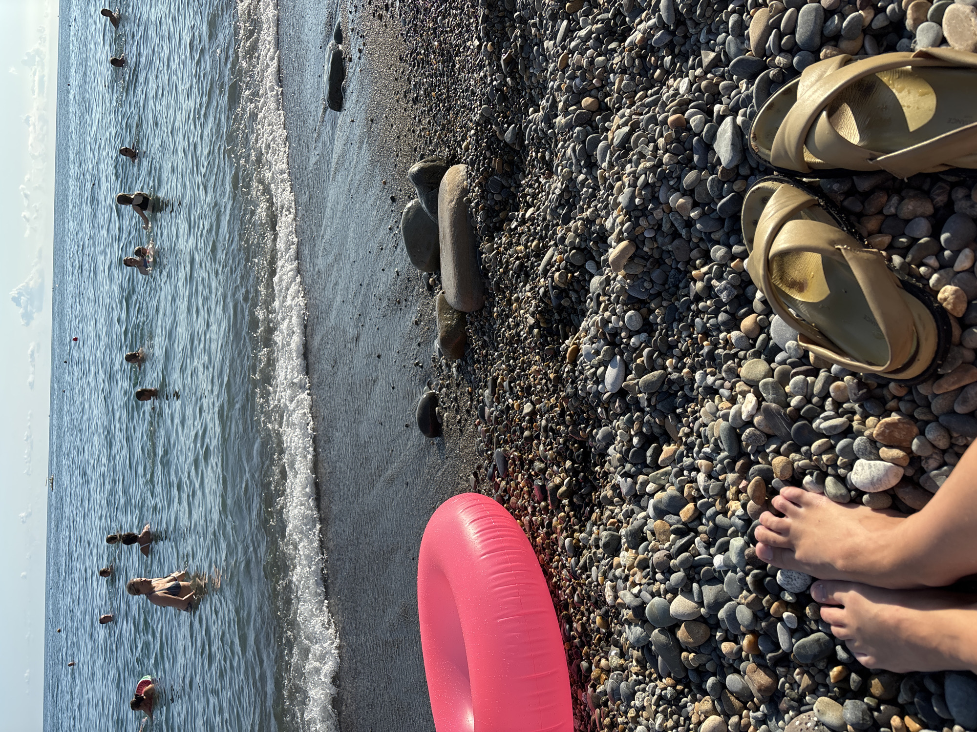 A beach scene on a sunny day, likely in Sochi, Russia, features a pebbled shoreline with a bright pink inflatable ring resting on the stones. The sea, with gentle waves breaking on the shore, is populated by numerous people swimming and enjoying the water. In the foreground, a person's bare feet and a pair of beige sandals are visible on the rocky beach. The sky is clear with a few scattered clouds.