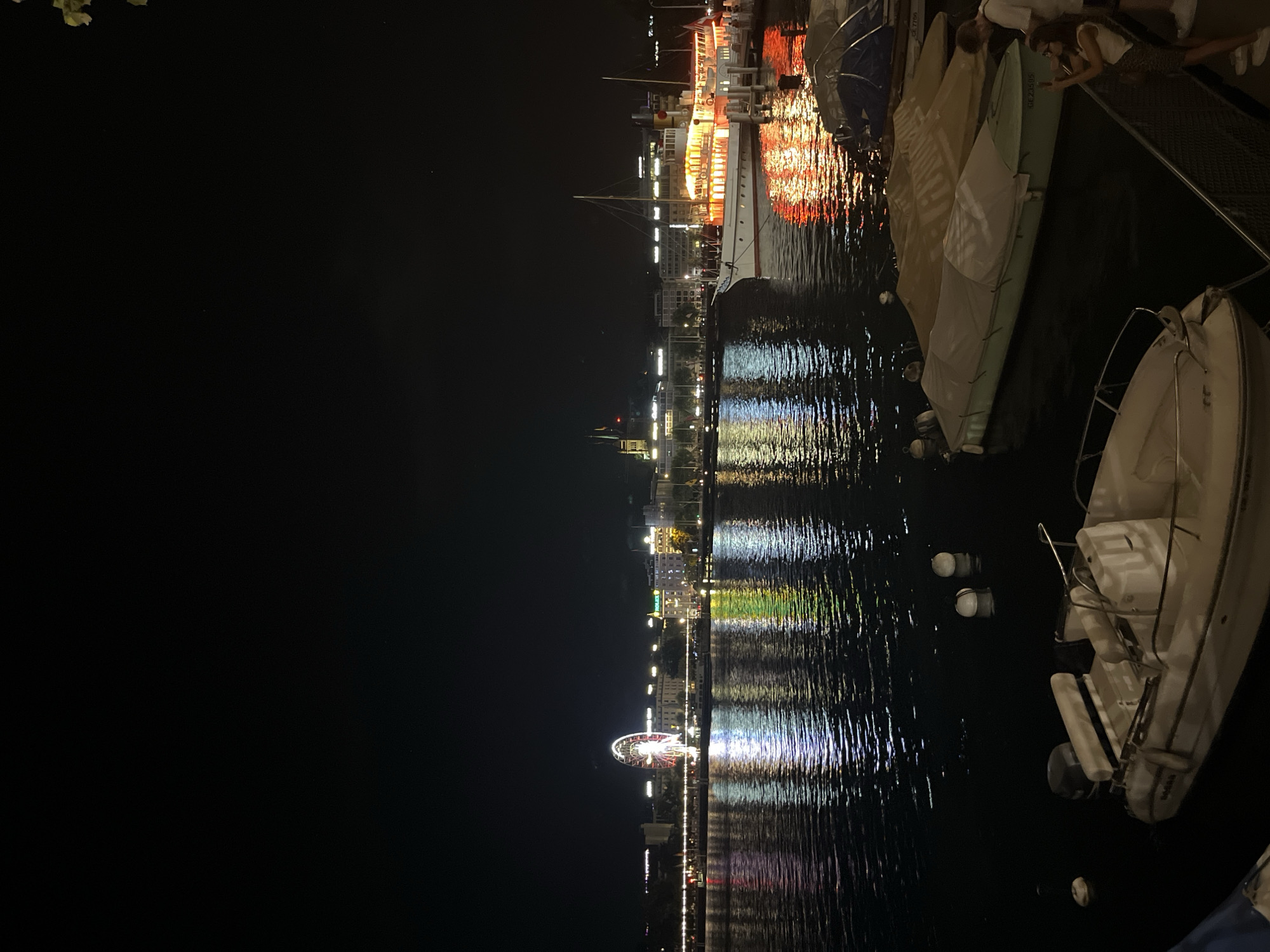 The image depicts a nighttime scene at the Jet d'Eau in Geneva, Switzerland. The Ferris wheel, illuminated with colorful lights, stands prominently in the background, reflecting on the calm waters of Lake Geneva. Several boats are moored in the foreground, some covered with tarps. A few people are visible on the right side of the image, one of whom appears to be taking a photograph of the cityscape. The surrounding buildings are also lit up, creating a vibrant and festive atmosphere.
