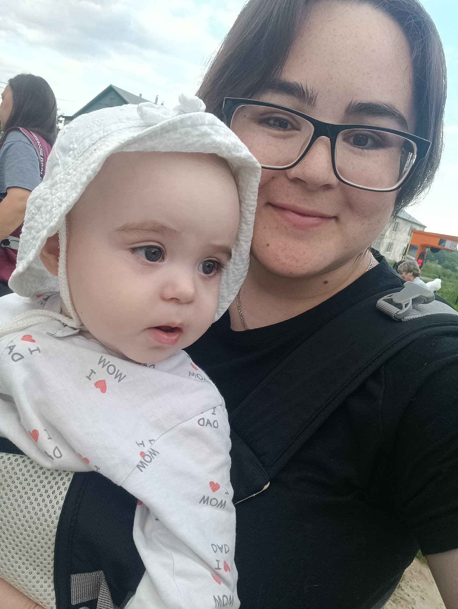 A close-up selfie features a woman and a baby in a carrier. The woman, wearing glasses and a black shirt, is on the right side of the frame, holding the baby on her left. The baby, wearing a white sun hat and a white onesie with "MOM" printed on it, is to the left of the woman. The background shows a building, trees, and a cloudy sky. The location is not identifiable in the city of Посад, Russia, from the image.