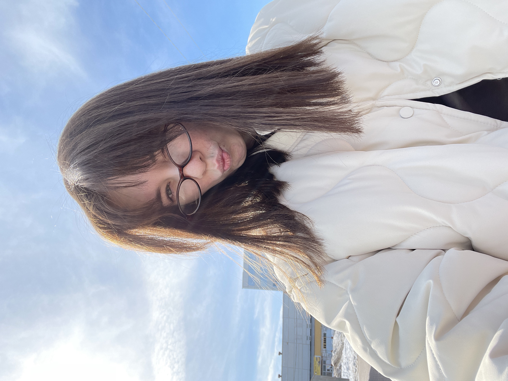 The image is a selfie of a young woman with brown hair and glasses, wearing a white quilted jacket. She is looking directly at the camera with a slightly puckered expression. The background features a blue sky with scattered clouds and a building in the distance. Based on the architecture and signage visible in the image, the location is identified as Chaikovsky, Russia. The photograph appears to be taken outdoors during daylight hours.