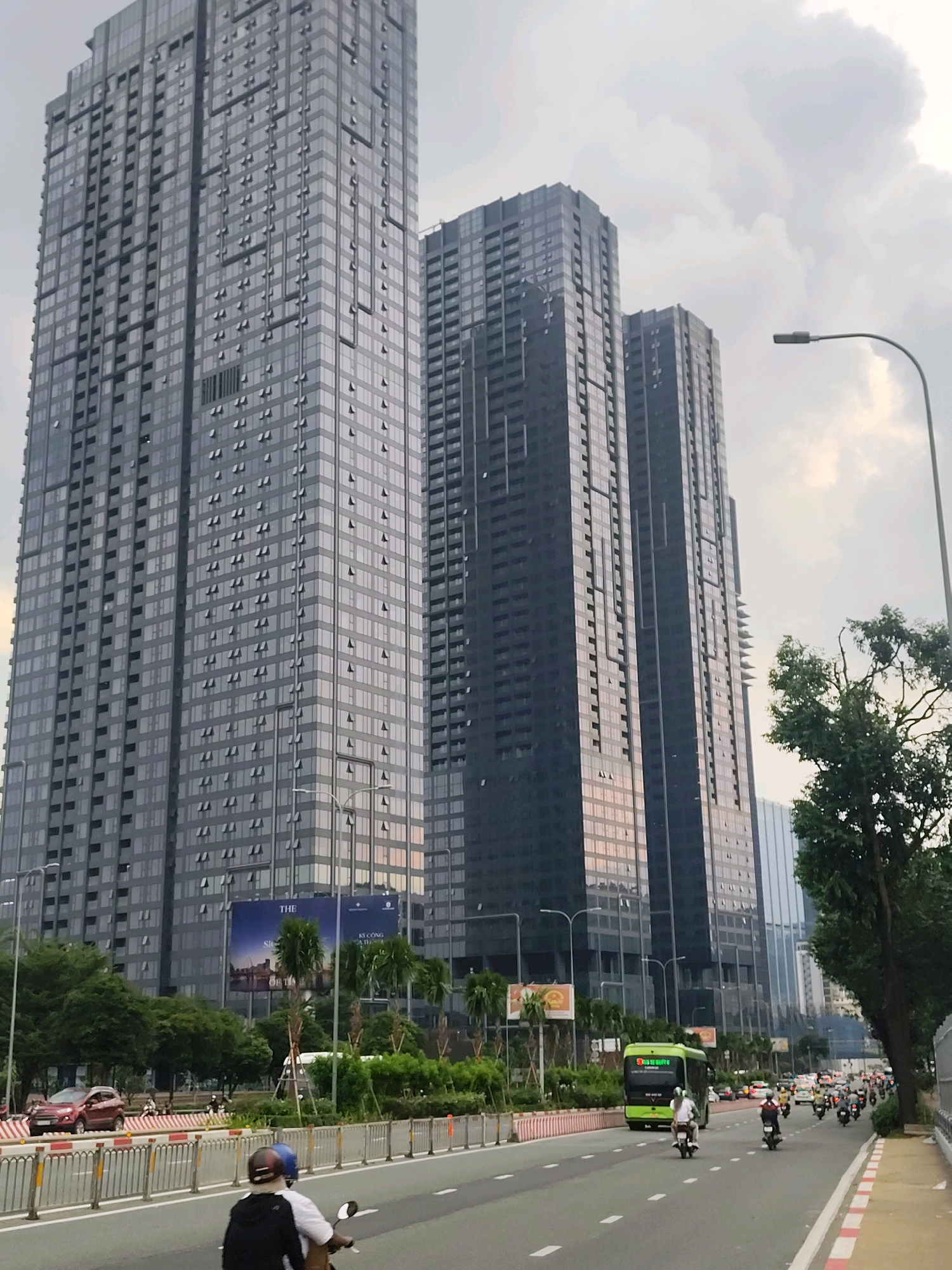 In Ho Chi Minh City, Vietnam, a wide, multi-lane road runs alongside several towering, modern skyscrapers with reflective glass facades. The sky is overcast with dramatic clouds. On the road, traffic moves in both directions. A green bus with "ECO BUS" written on its side is in the right lane, followed by several motorbikes. In the left lane, a red car is visible, also in traffic. On the pavement, planters with palm trees and other greenery are interspersed with large billboards, one of which reads "THE". A streetlamp with a curved arm stands tall on the right side of the road. The foreground features a close-up of a person riding a motorbike, wearing a helmet and a black and white jacket. The scene depicts a typical urban environment in a bustling Vietnamese city, characterized by high-rise architecture and active street life.
