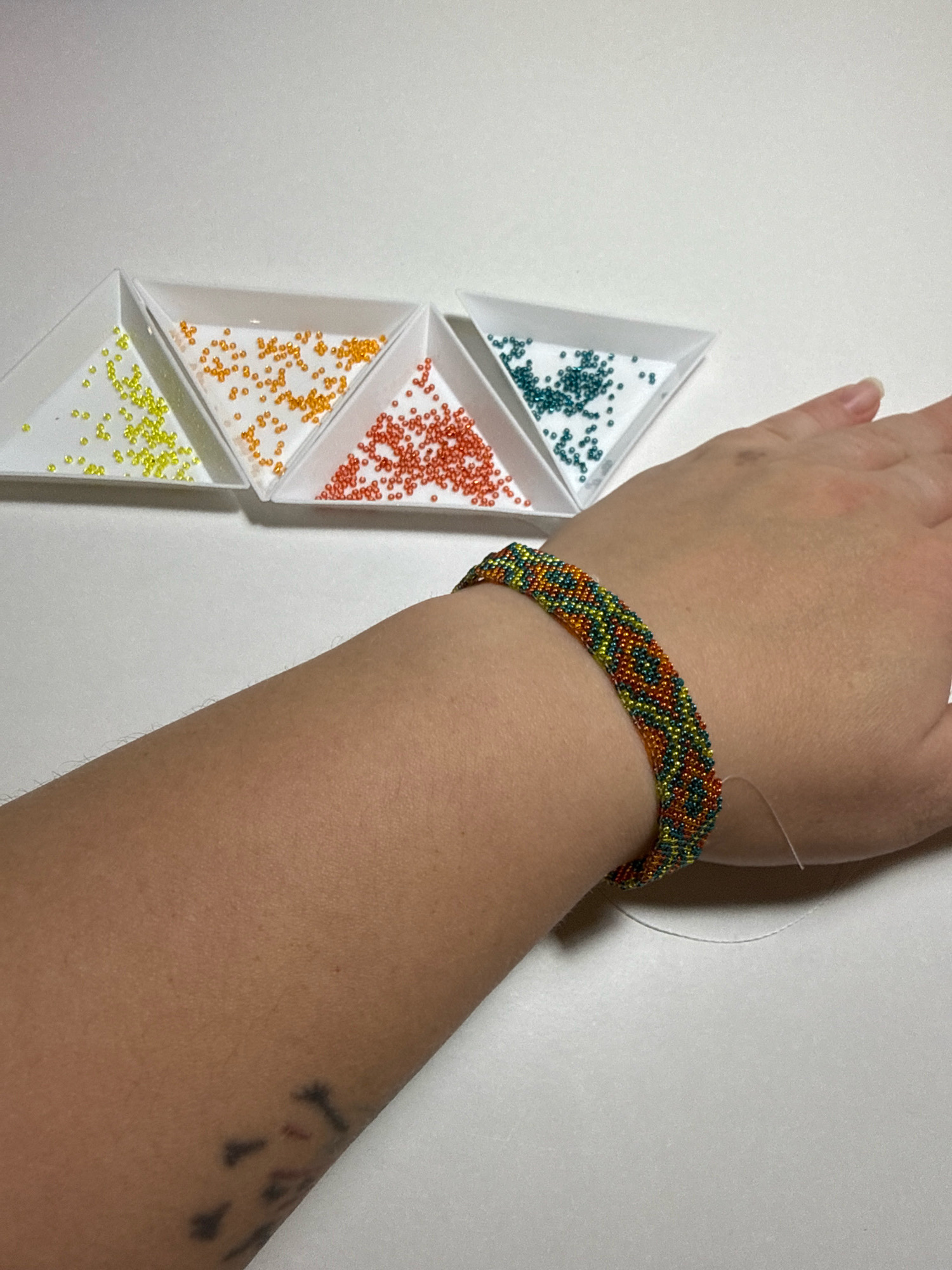A person's forearm and hand are visible, wearing a multi-colored beaded bracelet with a pattern of orange, green, yellow, and teal beads. In the background, four triangular dishes of small beads are arranged in a row. The dish on the far left contains yellow beads, the next dish has orange beads, followed by a dish of red beads, and finally, a dish of teal beads. The background is a plain white surface. There is no indication of the location being Kazan, Russia.