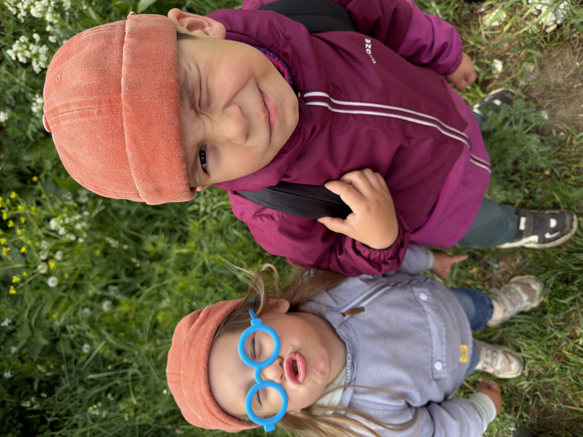 This overhead image captures two young children enjoying a playful moment outdoors in Chaikovsky, Russia. Both children are positioned on a lush green lawn, dotted with small white and yellow wildflowers, looking upwards towards the camera.
The child in the upper part of the frame is wearing an orange beanie and a maroon or purple hooded jacket featuring white stripes and a visible "QUECHUA" brand logo on the chest. This child has dark hair, is winking with one eye, and smiling broadly, conveying a cheerful and mischievous expression.
Below them, another child is also wearing an orange beanie and a light grey or blue zip-up jacket over blue jeans. This child has long brown hair flowing around their head and is making a humorous "fish face" with puckered lips, while playfully wearing bright blue, round-framed novelty glasses without lenses. Both children are wearing sneakers visible on the grass.
The scene depicts a joyful and casual outdoor activity, likely a family outing or playtime in a natural setting during the daytime. The expressions of the children suggest a lighthearted and fun atmosphere.