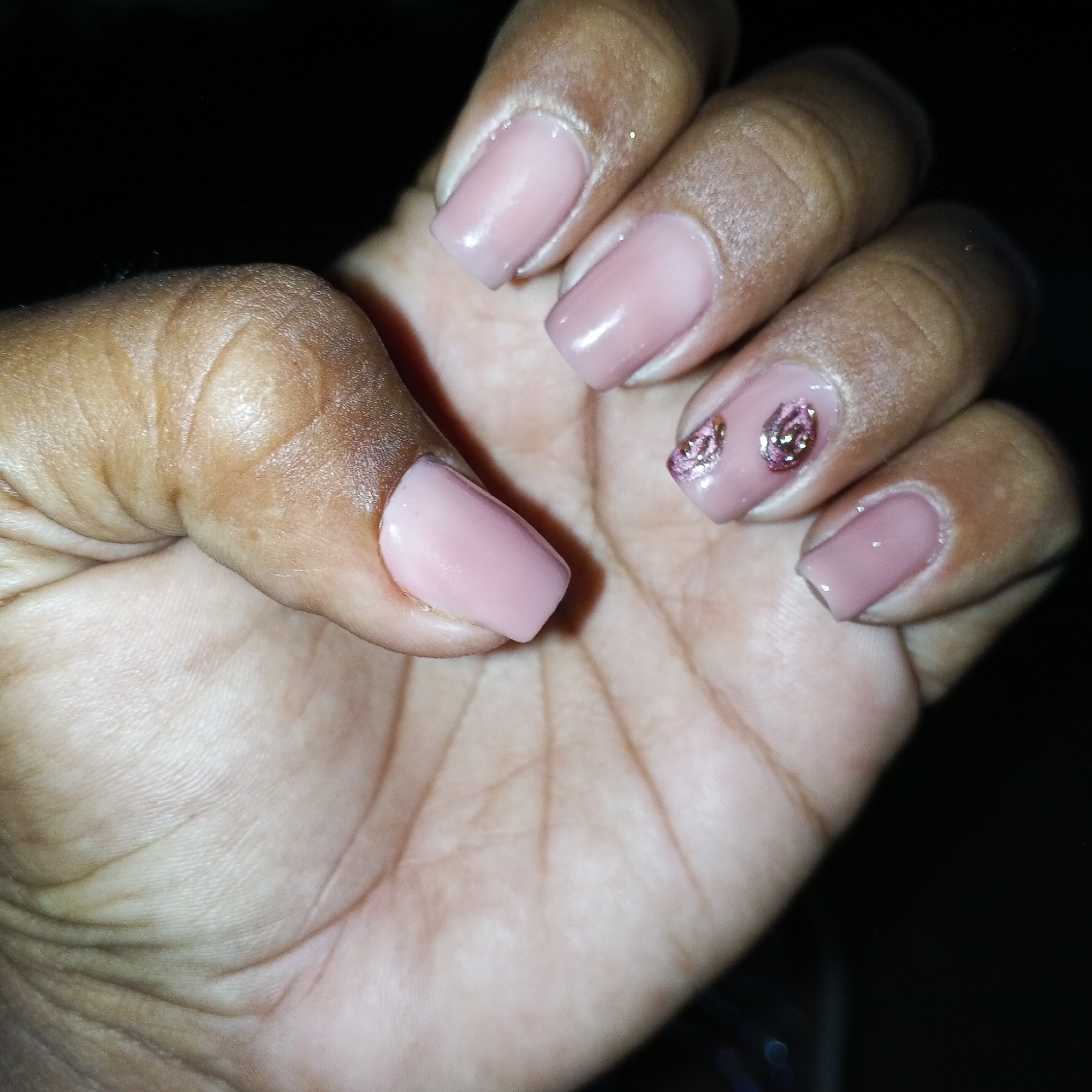 A close-up image shows a dark-skinned hand with manicured fingernails. The nails are painted a light, dusky rose color, with one nail on the right hand adorned with glittery embellishments in two small circular patterns. The lighting appears to be dim, with shadows visible on the hand and in the background, which is dark and undefined. The skin on the hand appears slightly dry.
