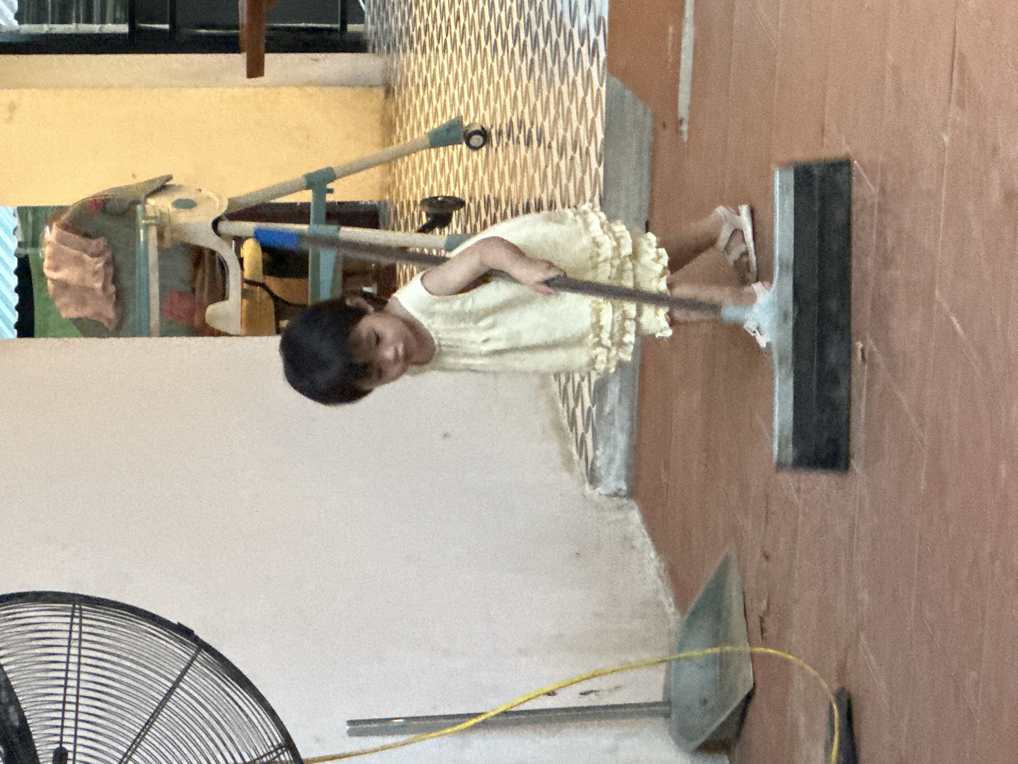 A young child, appearing to be a toddler, is sweeping a tiled floor with a broom. The child is wearing a light-colored, ruffled dress and sandals. The setting appears to be an outdoor or semi-outdoor area, possibly a patio or courtyard, with a tiled floor and a wall in the background. A large fan is visible on the left side of the image, and a dustpan is on the floor near the child. In the background, a high chair with blankets draped over it is visible, suggesting a domestic setting. The overall scene is one of a child engaged in household chores.