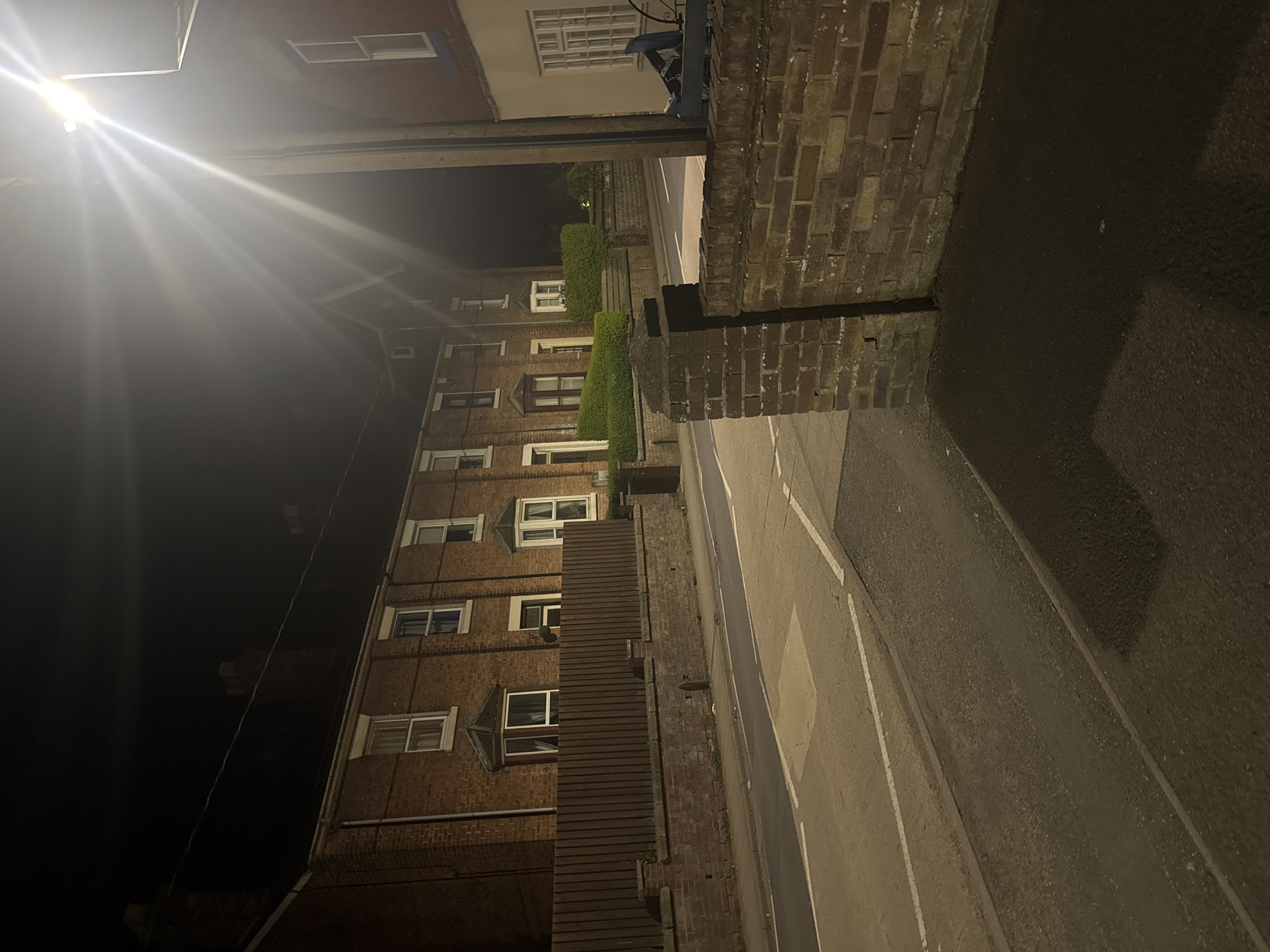 The image depicts a nighttime street scene in Welshpool, United Kingdom. A row of brick buildings with multiple windows is visible along the left side of the frame. A street lamp positioned at the top left casts a strong glare, illuminating the buildings and street. The road surface is visible on the right side, marked with white lines. A brick wall runs parallel to the street. No people are visible in the frame. The environment is urban and lit by artificial light sources.